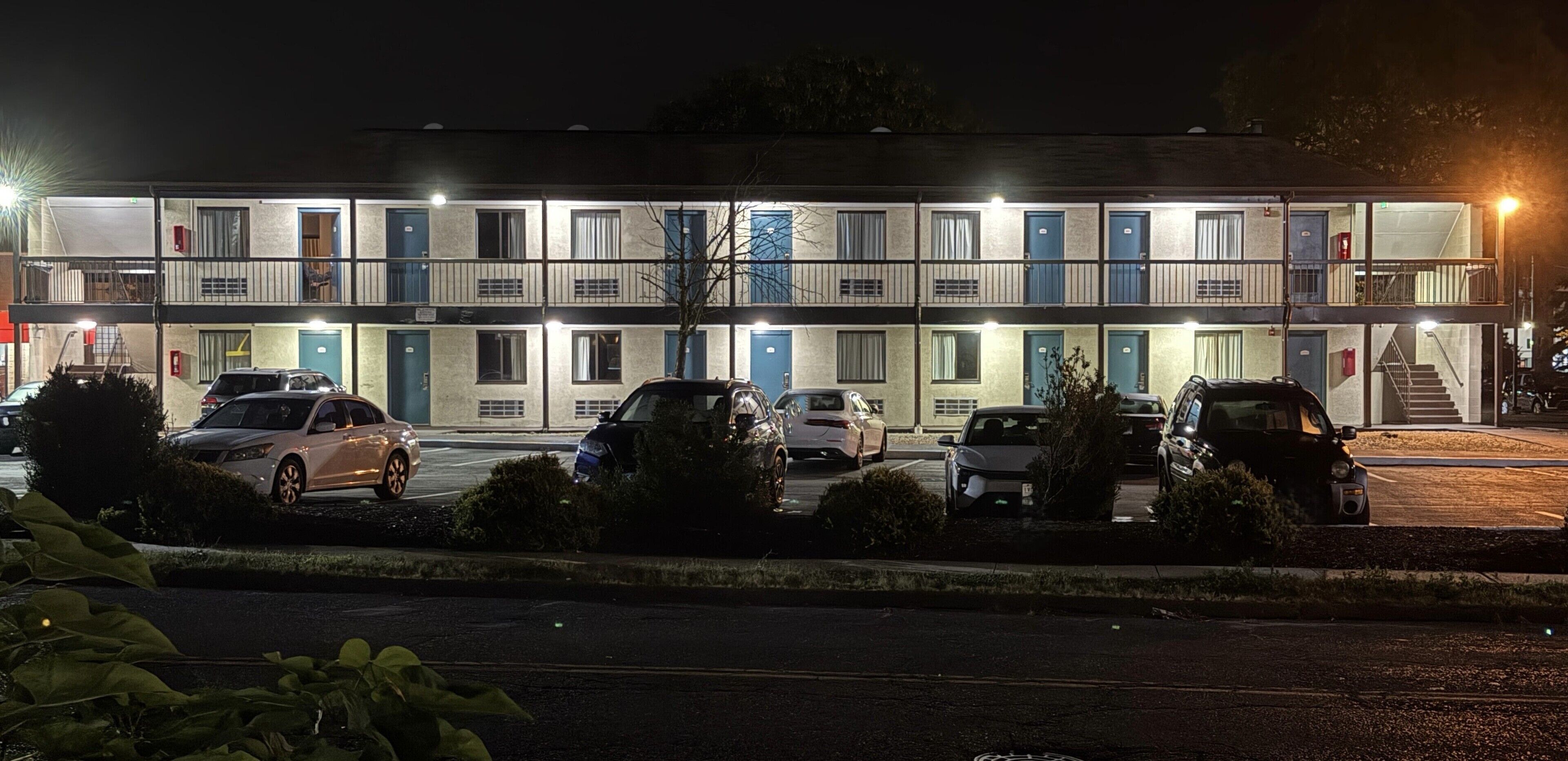 Un motel à étages, avec une rangée de chambres à chaque étage, un parking où sont garées plusieurs voitures et un escalier menant aux étages supérieurs.