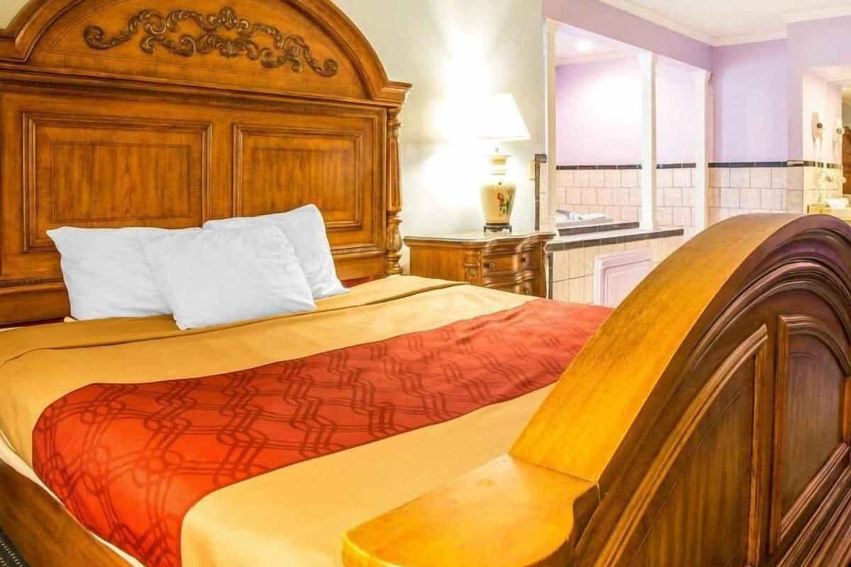 A hotel room with a wooden headboard, a bed with a patterned bedspread, a nightstand with a lamp, and a view of the bathroom through a glass door.