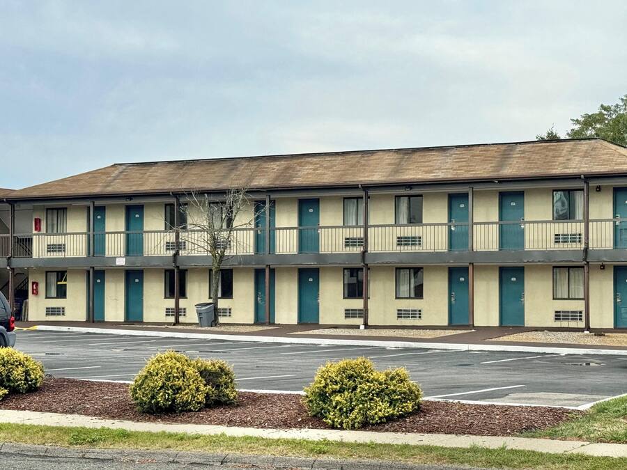 A motel with a row of rooms, a parking lot, and small bushes in front.