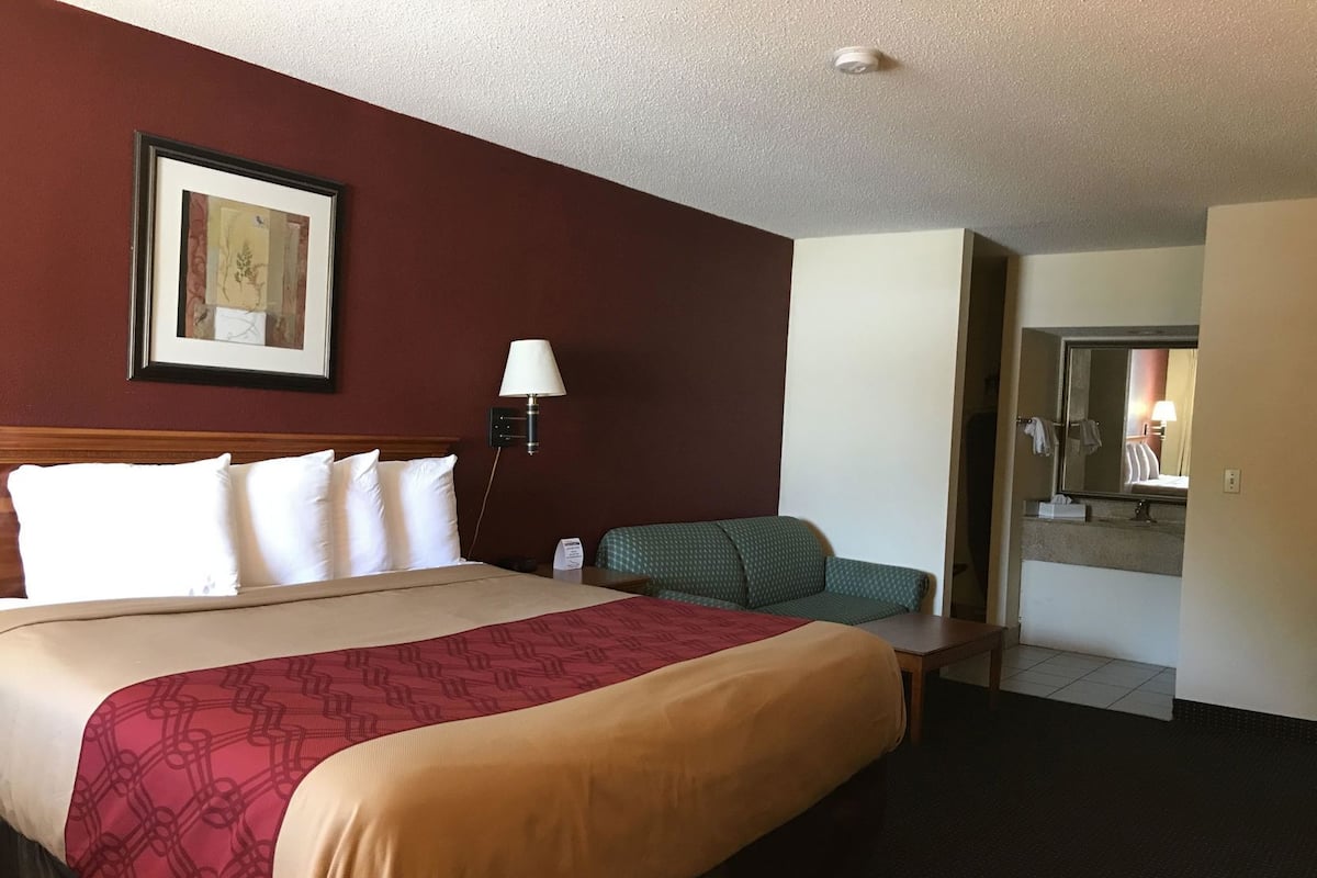 A hotel room with a large bed, a bedside table, a lamp, a green chair, and a bathroom visible through an open door.
