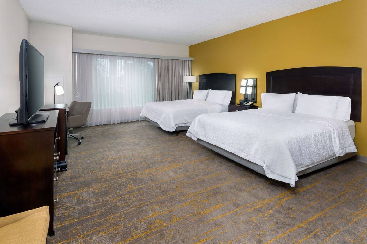 A hotel room with two beds, a desk with a TV, and a large window with curtains.