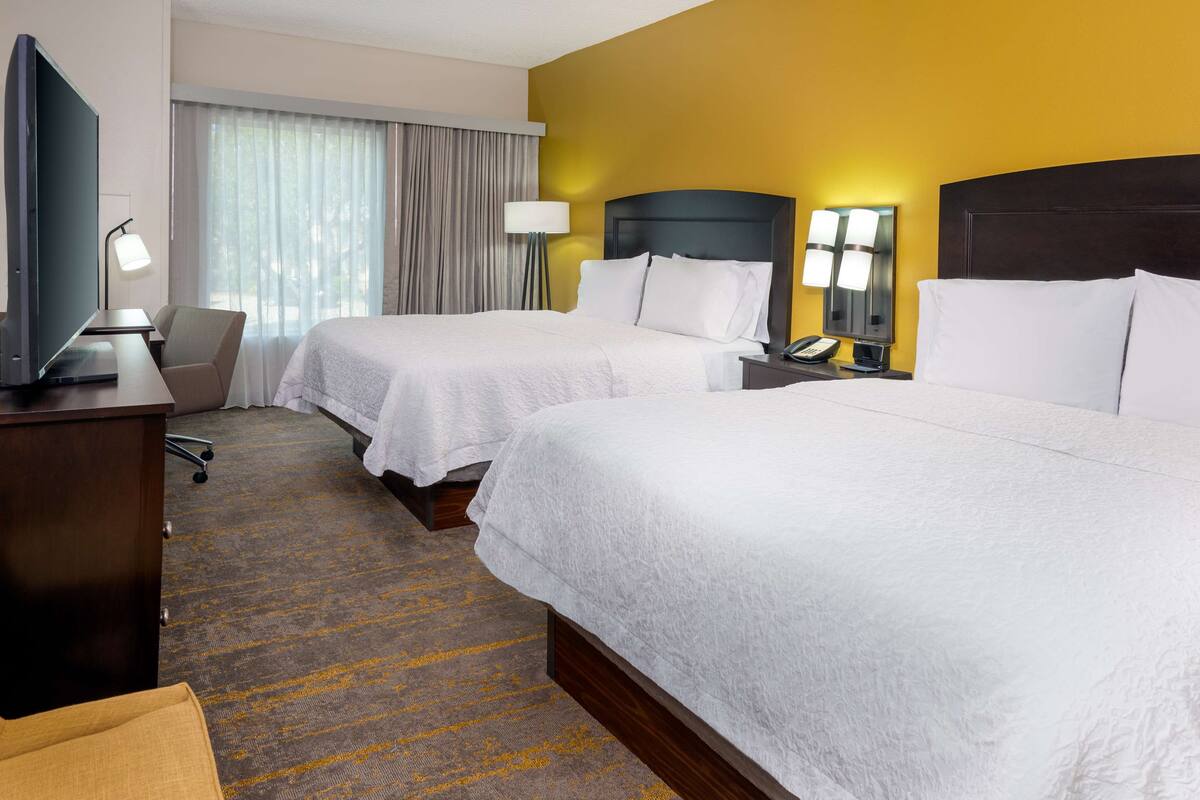 A hotel room with two beds, a desk with a computer, a chair, a lamp, and a window with curtains.