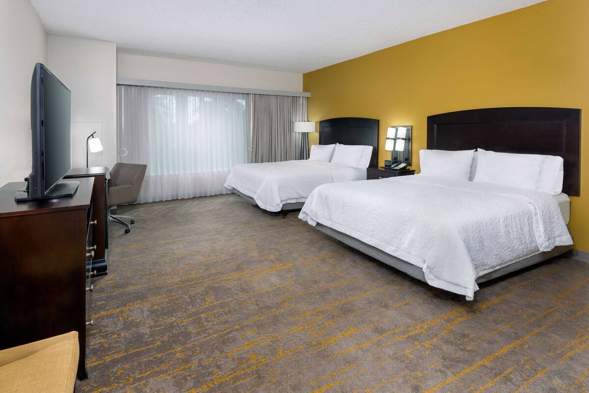 A hotel room with two beds, a desk with a TV, a chair, and a large window with curtains.