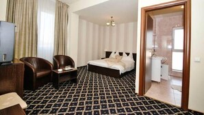 Habitación de hotel con cama, dos sillas, una mesita, televisión y baño a la vista a través de una puerta abierta.