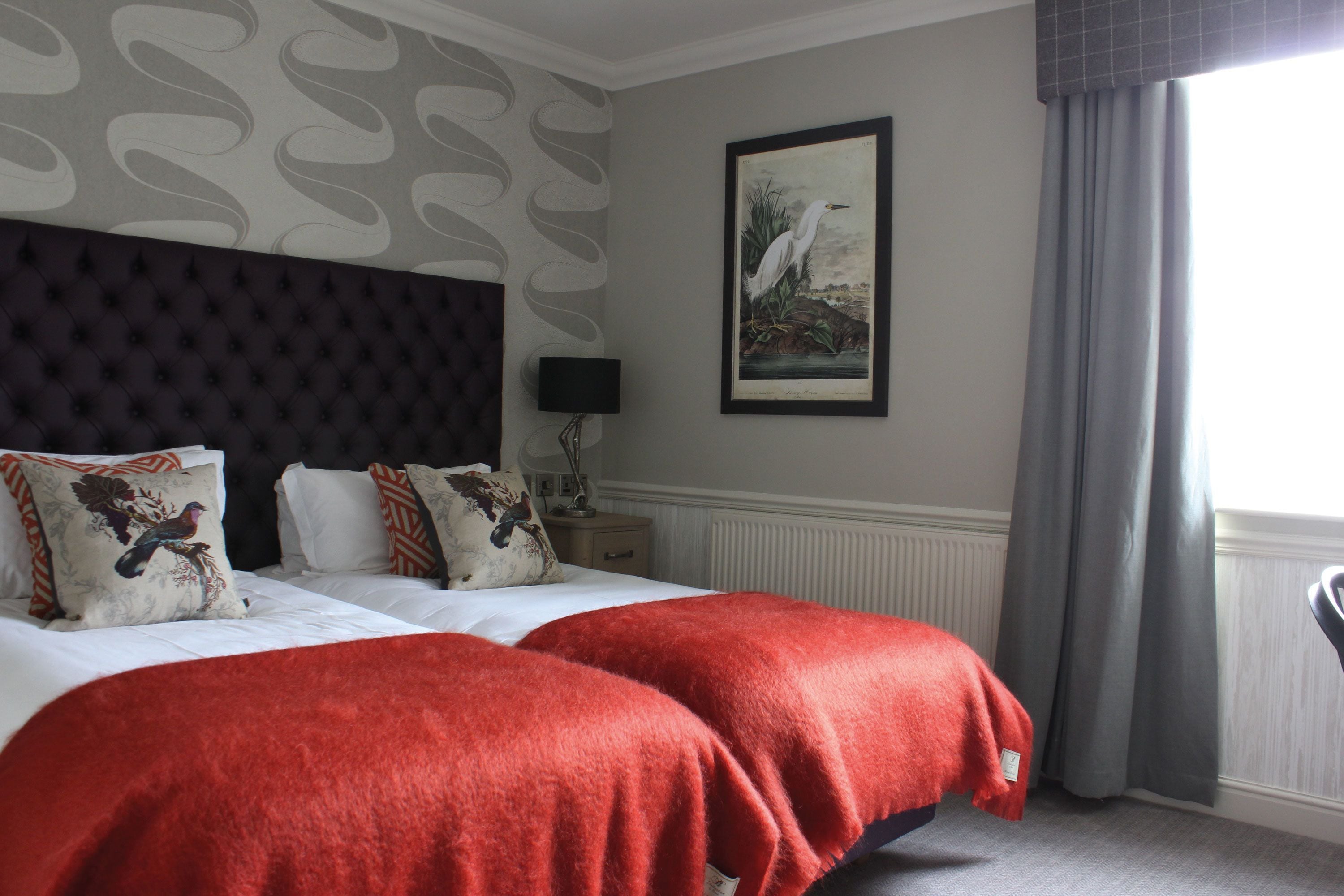 A hotel room with a large bed, two red throw blankets, a dark headboard, a framed painting of a bird, a nightstand with a lamp, and a window with curtains.