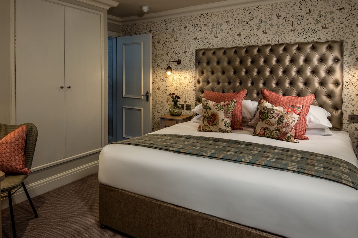 A bedroom with a tufted headboard, patterned pillows, a bedside table with a vase, and a door with floral wallpaper.