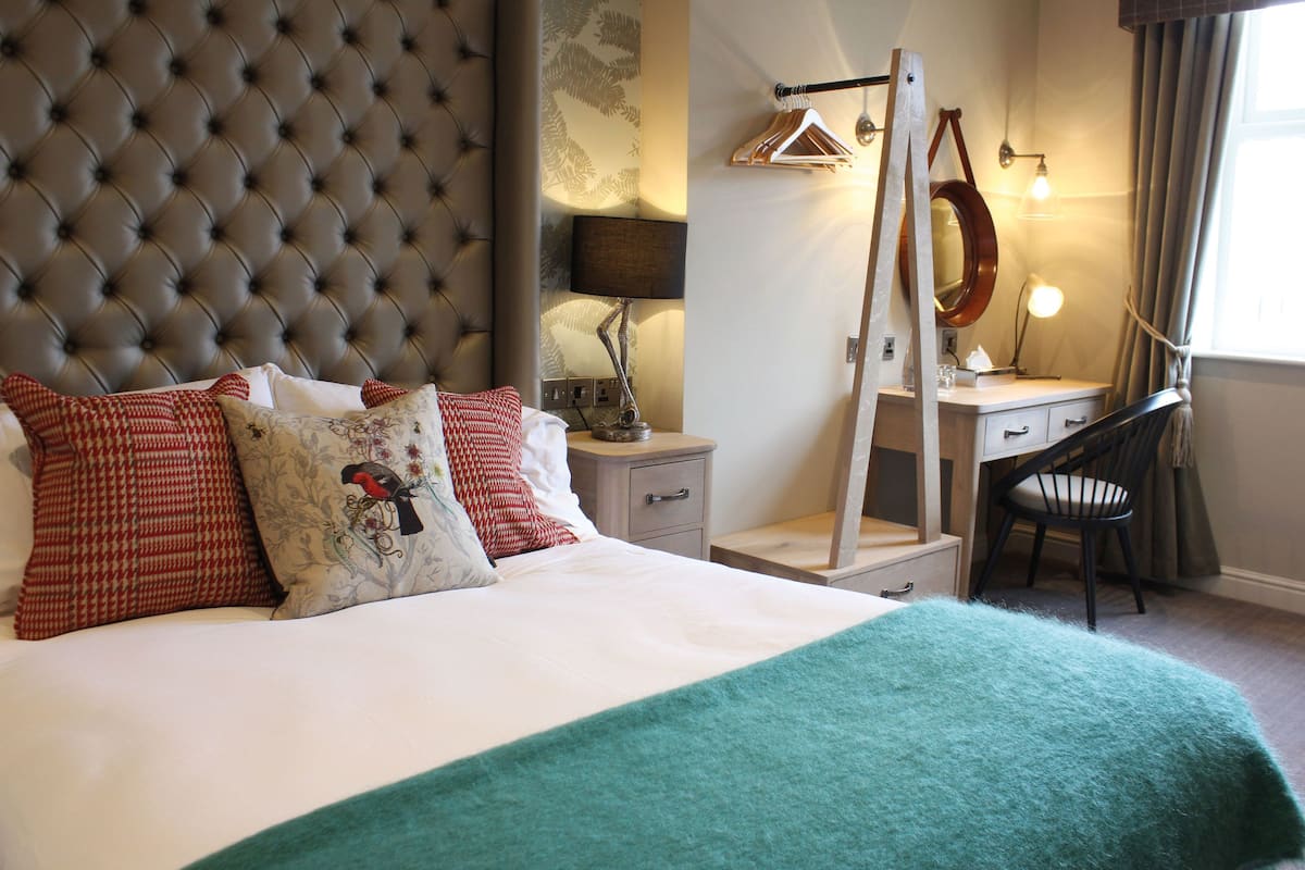 A bedroom with a tufted headboard, a bed with patterned pillows, a desk with a mirror, and a chair.