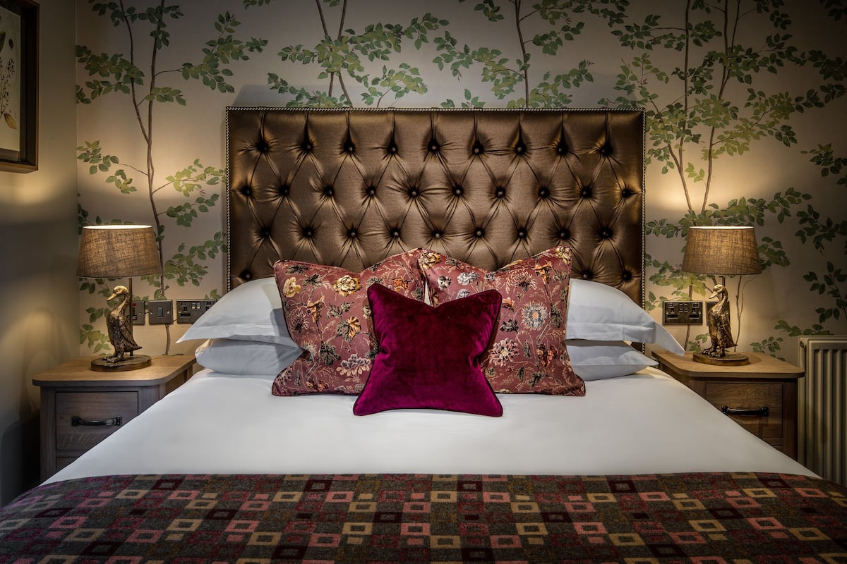 A neatly made bed with a patterned bedspread, a tufted headboard, and decorative pillows. There are two bedside tables with lamps and wall-mounted switches.