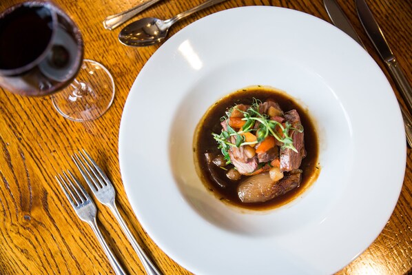 A white plate with a dish of meat and vegetables, a glass of red wine, and cutlery on a wooden table.