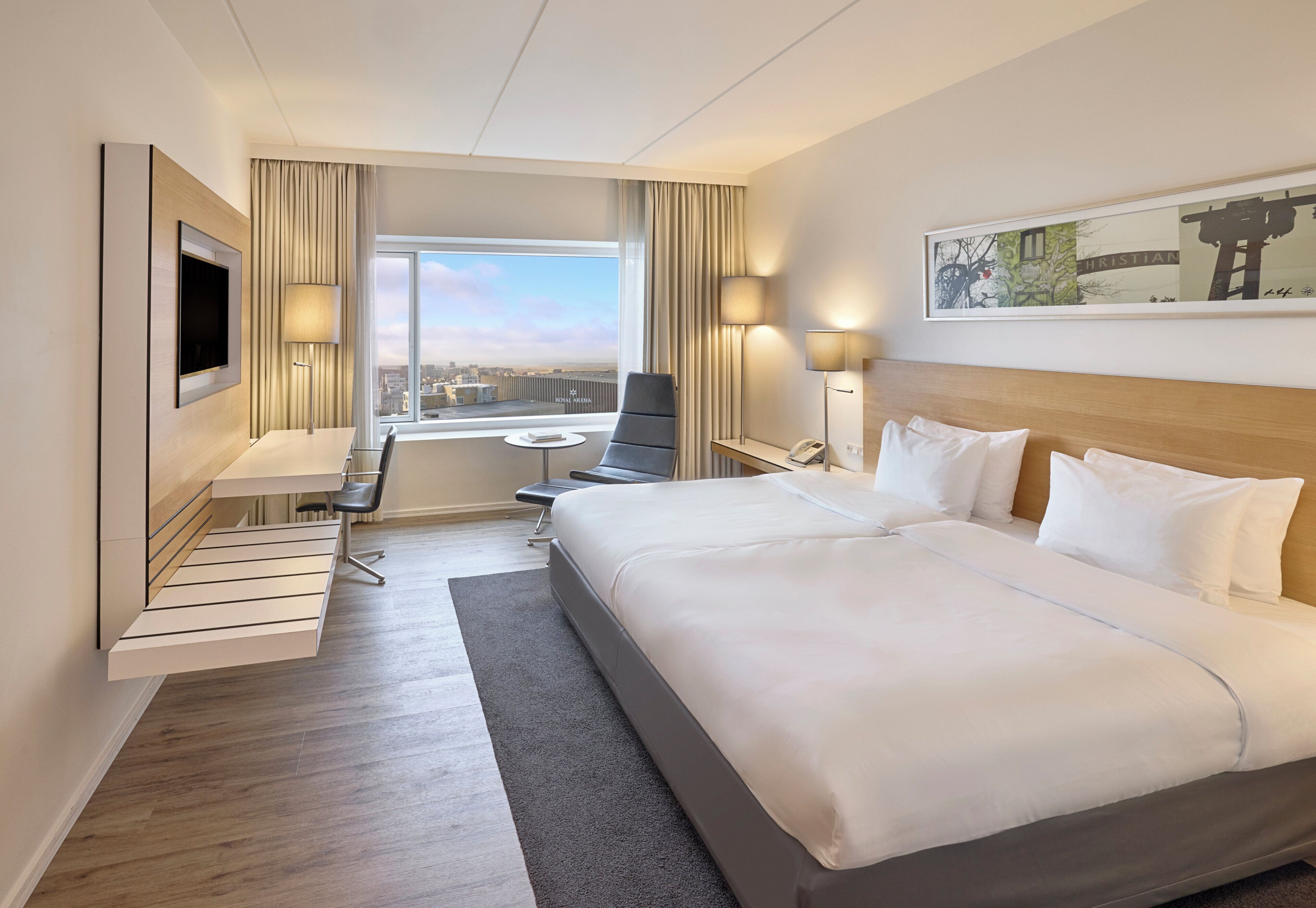 Ein Hotelzimmer mit einem großen Bett, einem Schreibtisch mit Stuhl, einem an der Wand befestigten Fernseher und einem Fenster mit Blick auf die Stadt.
