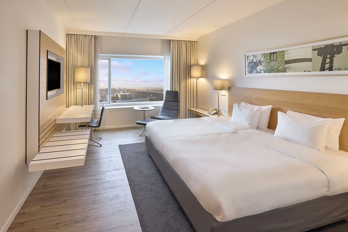 A hotel room with a large bed, a desk with a chair, a TV mounted on the wall, and a window offering a city view.