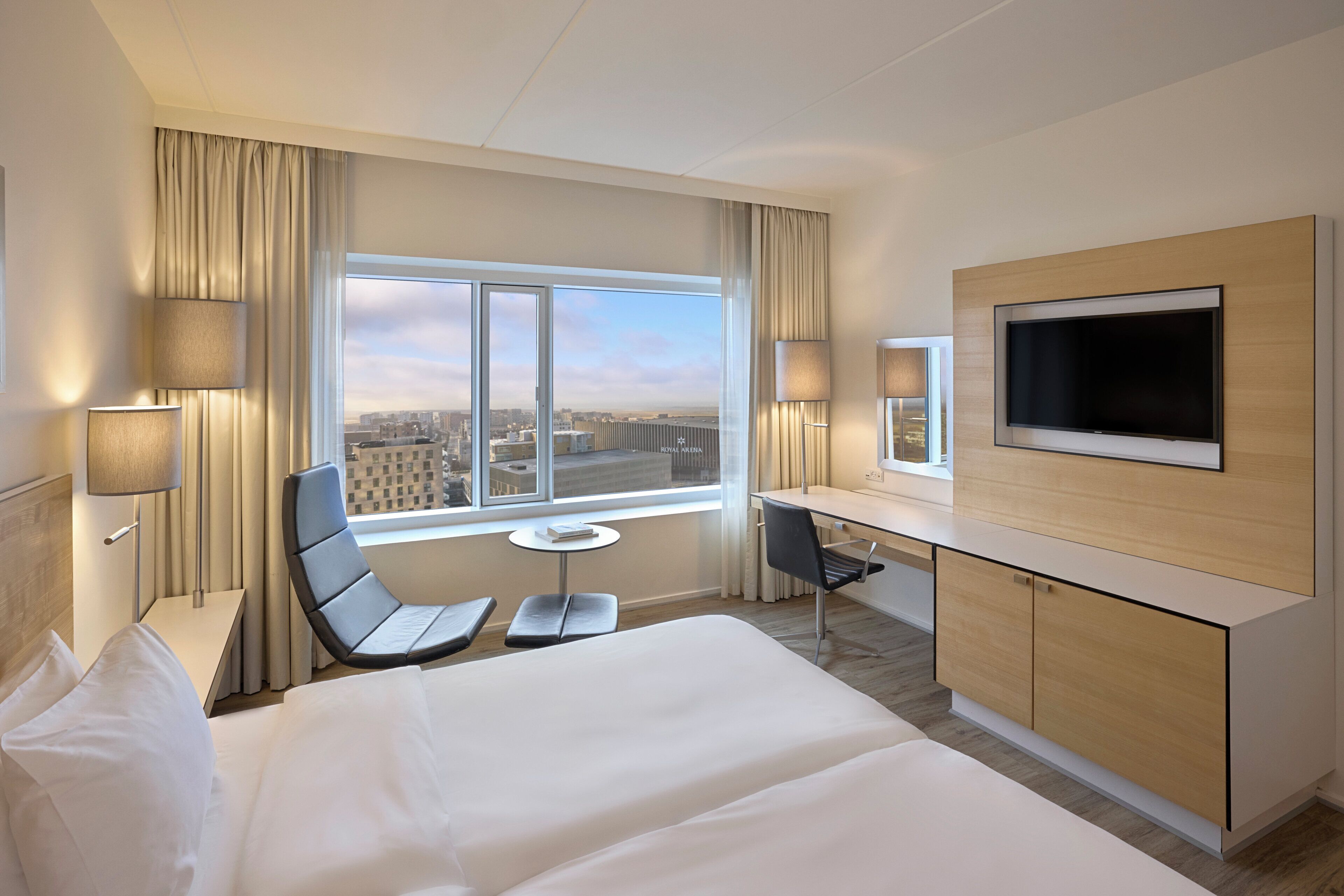 A hotel room with a bed, a desk, a chair, a TV, and a window with a city view.