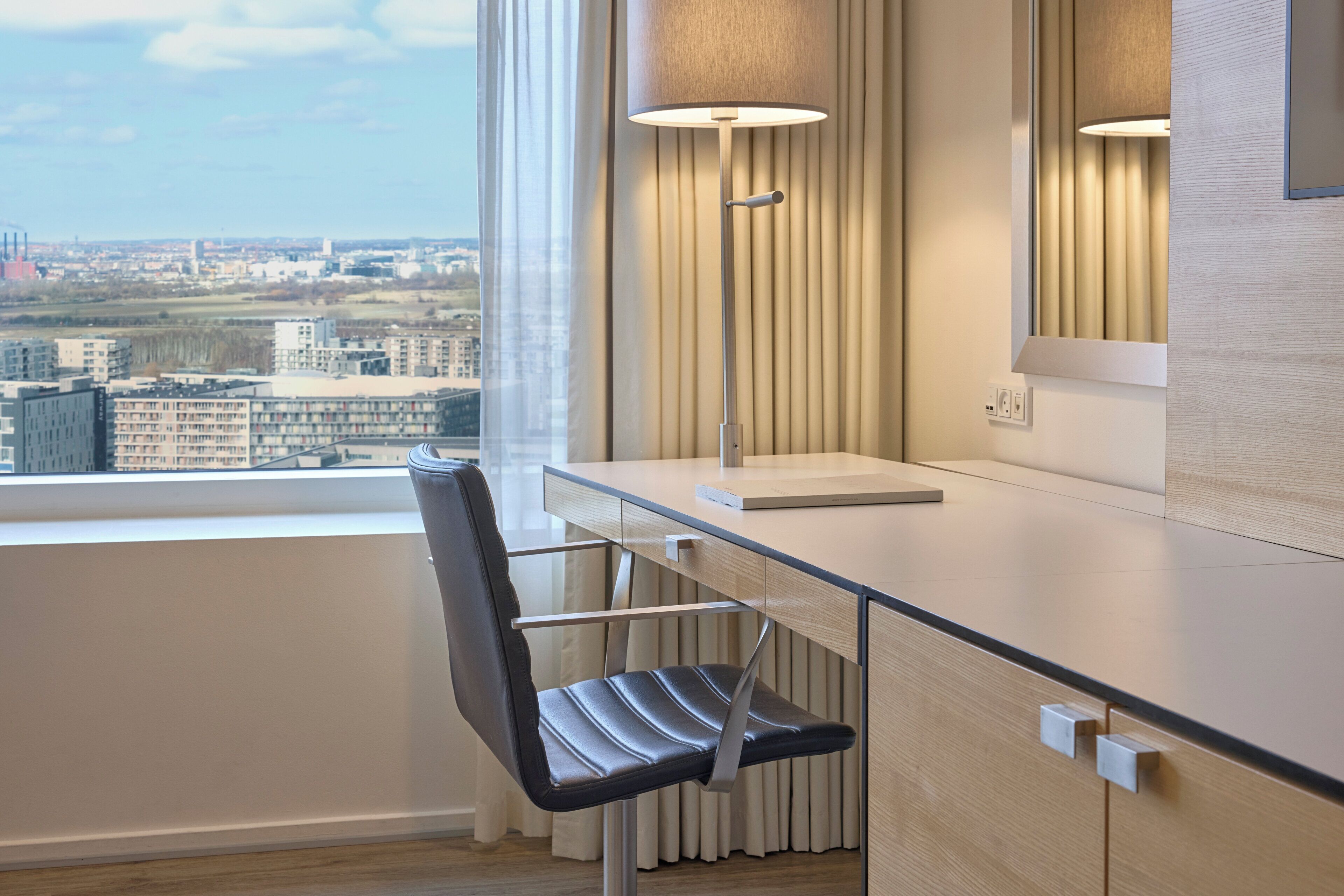 Une chambre d’hôtel moderne équipée d’un bureau, d’une chaise, d’une lampe et d’une grande fenêtre donnant sur la ville.