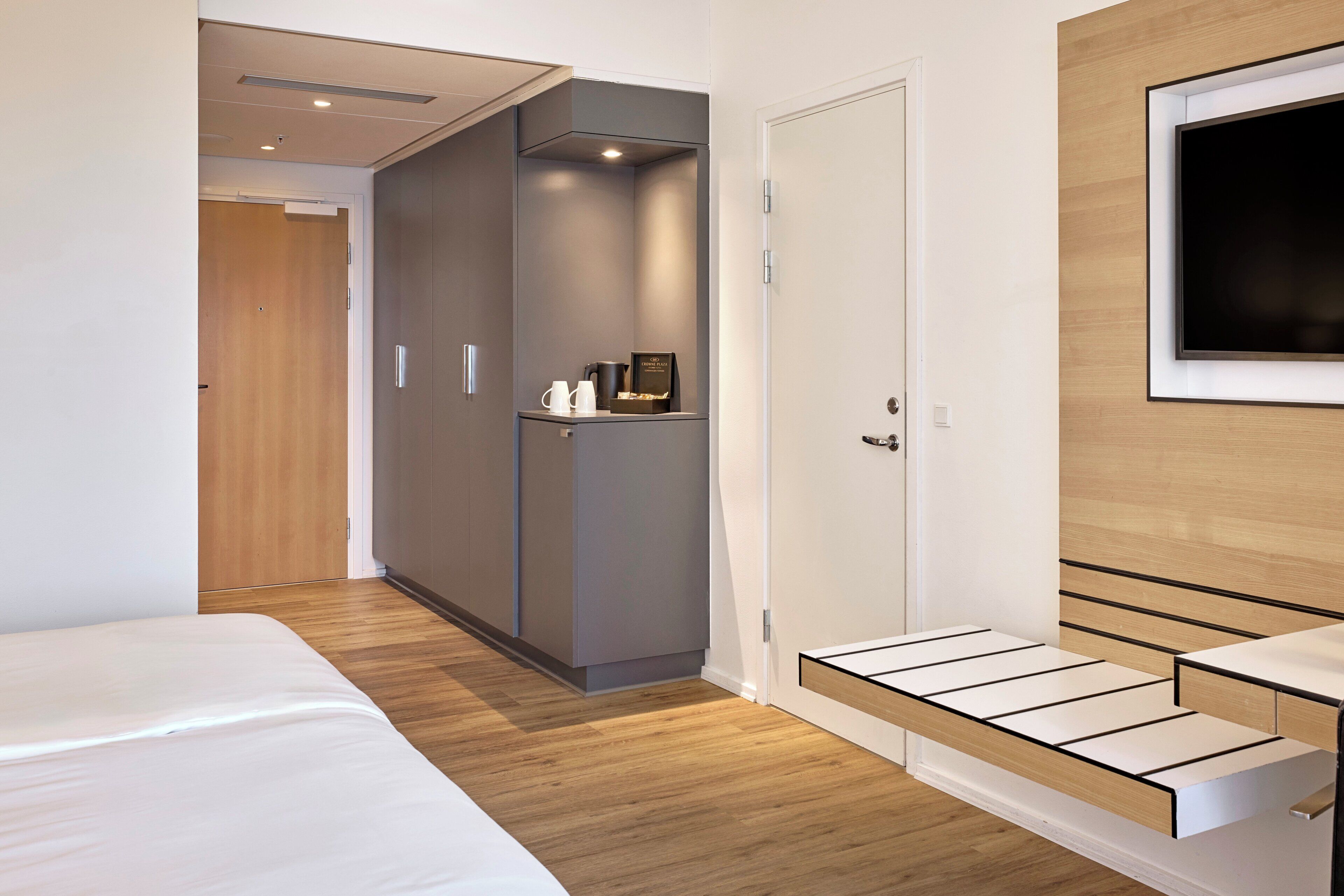 A modern hotel room with a bed, a wardrobe, a television mounted on the wall, and a small seating area.