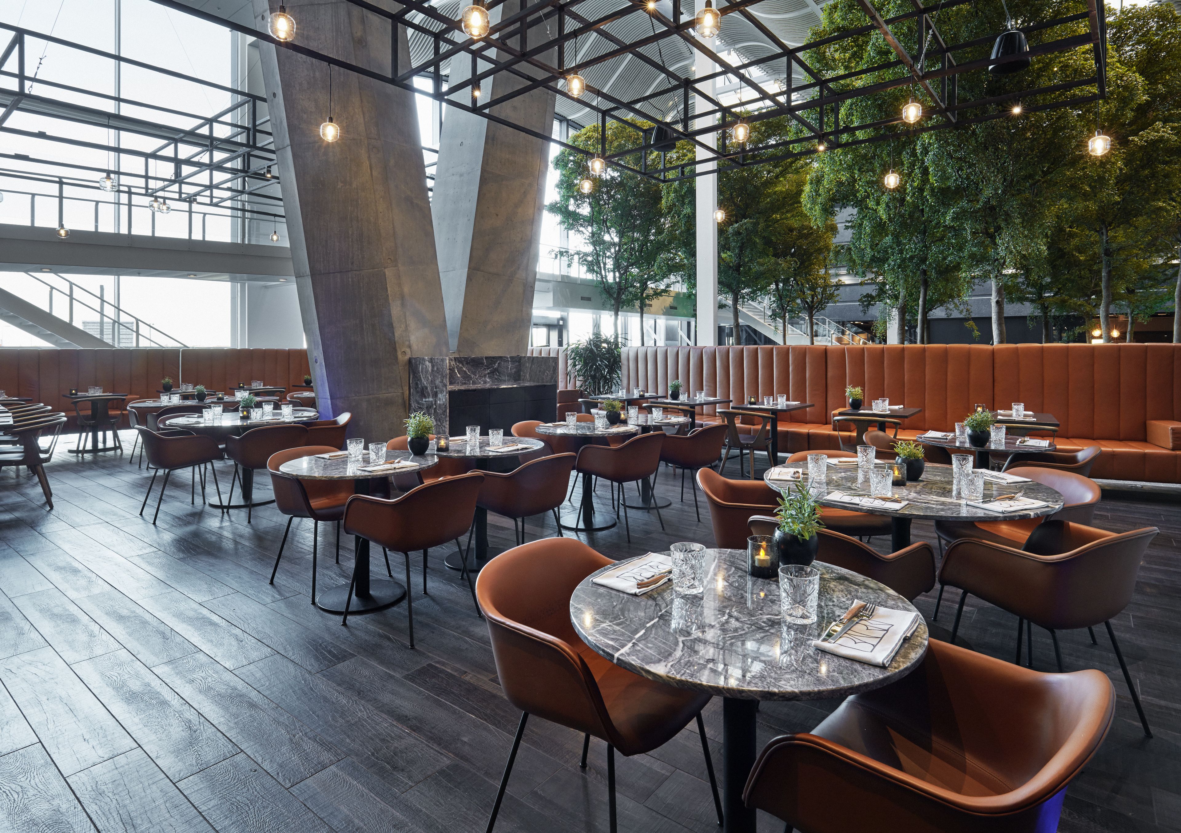 A modern restaurant with round tables and orange chairs, featuring a high ceiling and natural light from large windows.
