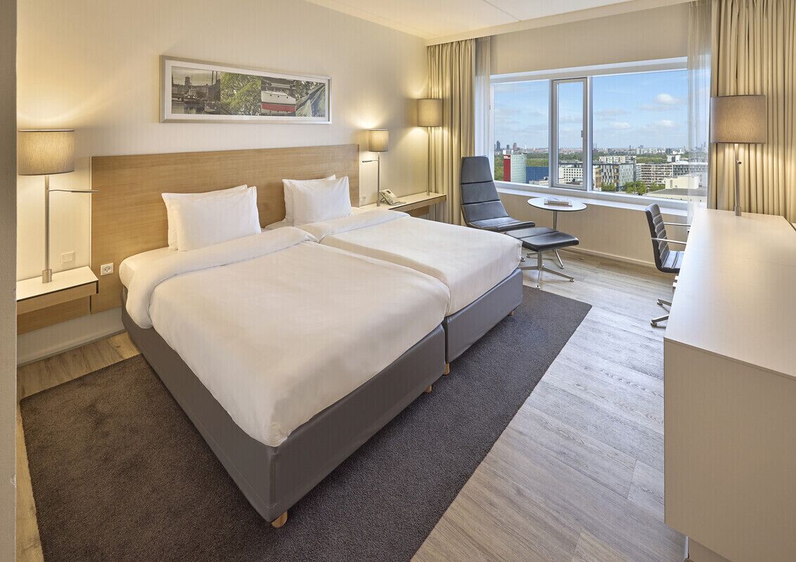 A hotel room with a large bed, a desk, a chair, and a view of the city.