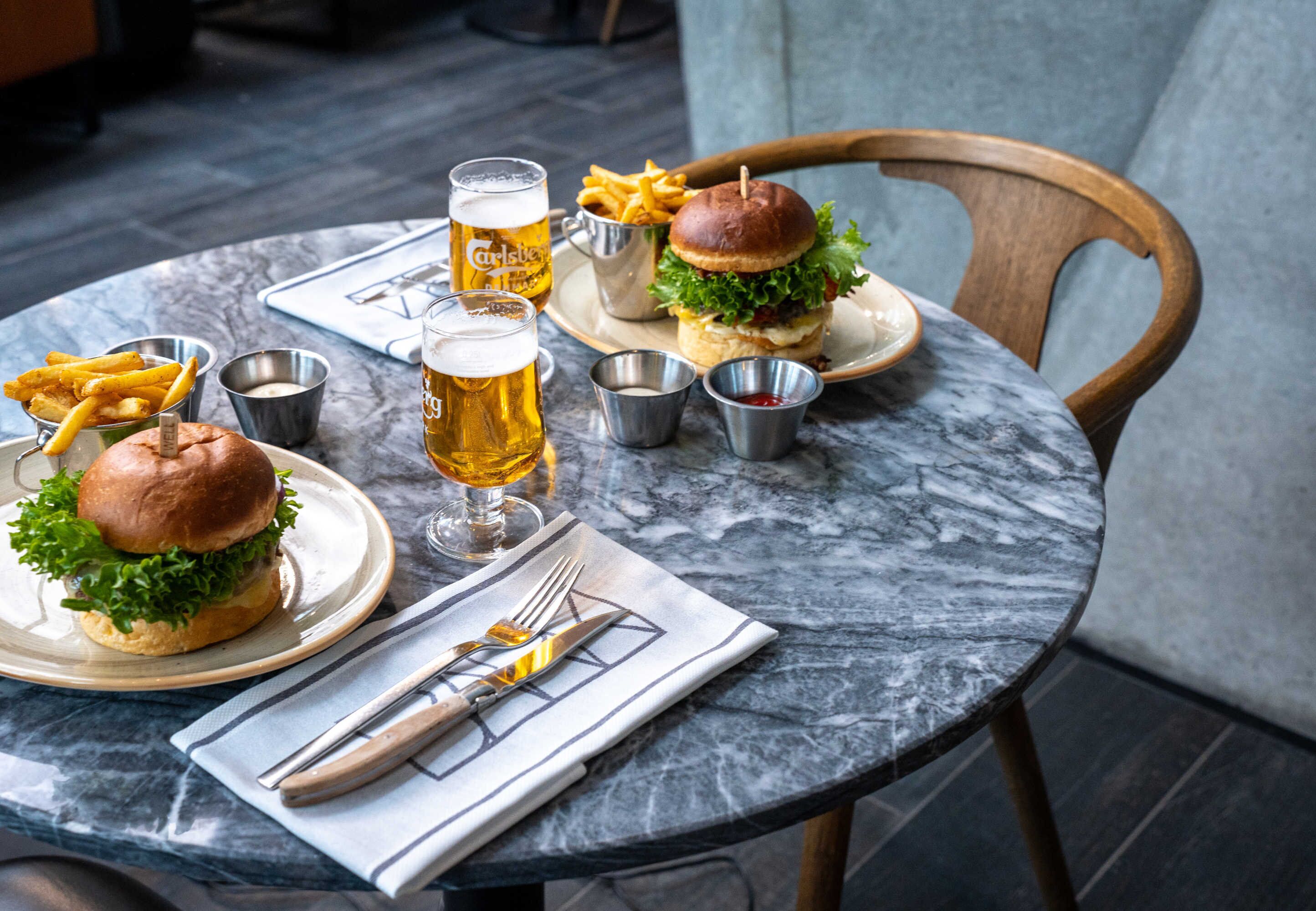 A dining table with two burgers, fries, and beer glasses.
