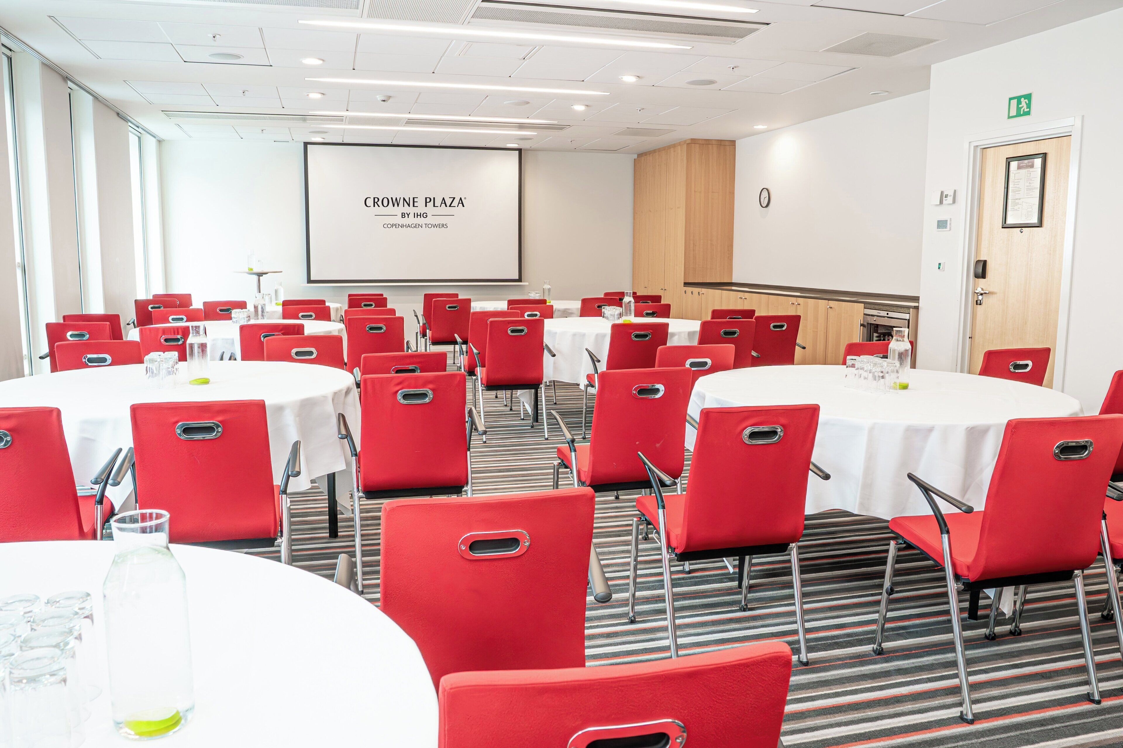 Ein Konferenzraum mit roten Stühlen und weißen Tischen, einer Projektionsleinwand, die 'CROWNE PLAZA BY IHG COPENHAGEN TOWERS' zeigt, sowie einem Holzkasten.