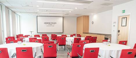 Ein Konferenzraum mit roten Stühlen und weißen Tischen, einer Projektionsleinwand, die 'CROWNE PLAZA BY IHG COPENHAGEN TOWERS' zeigt, sowie einem Holzkasten.