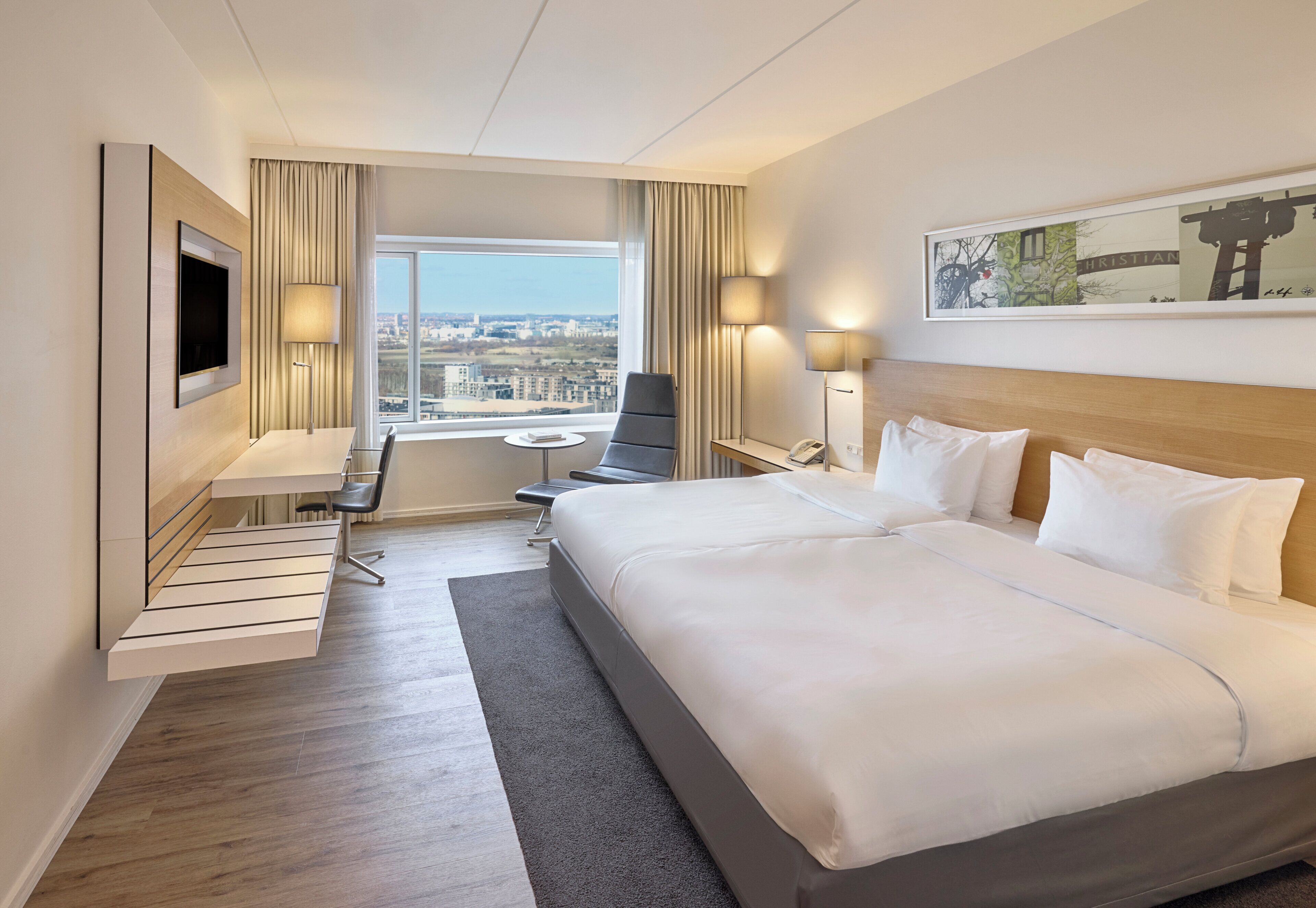 Ett hotellrum med en stor säng, ett skrivbord, en stol, en TV och ett fönster med utsikt över staden.