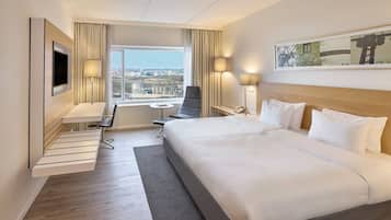 Ett hotellrum med en stor säng, ett skrivbord, en stol, en TV och ett fönster med utsikt över staden.