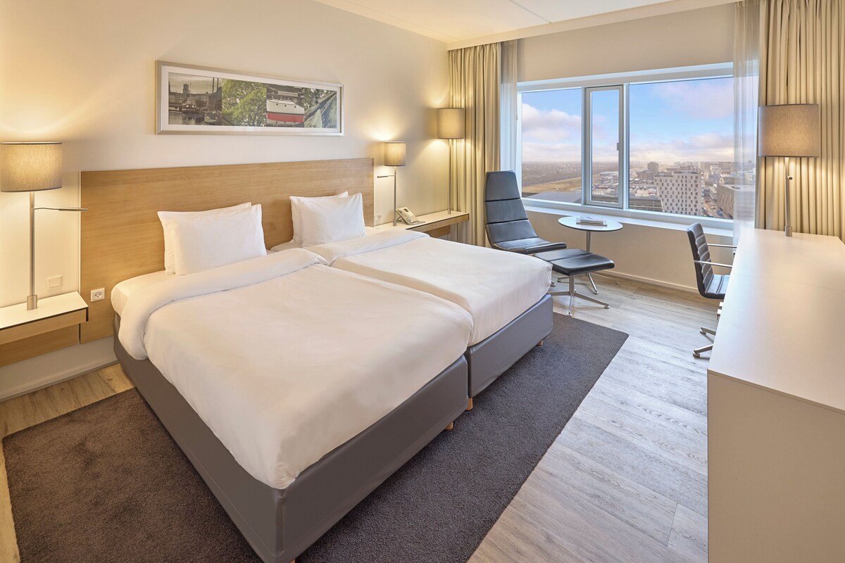 A hotel room with a large bed, a desk, a chair, and a view of the city.