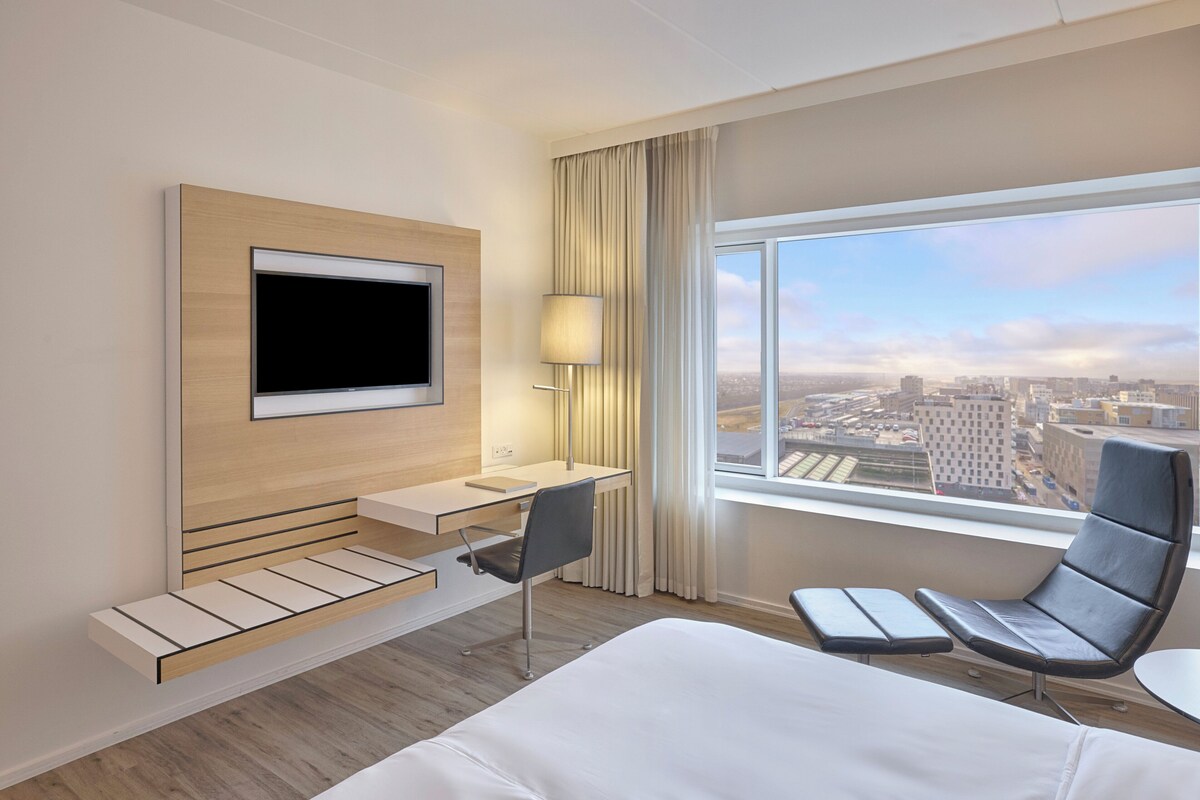 A modern hotel room with a bed, a desk, a chair, a TV, and a large window with a city view.