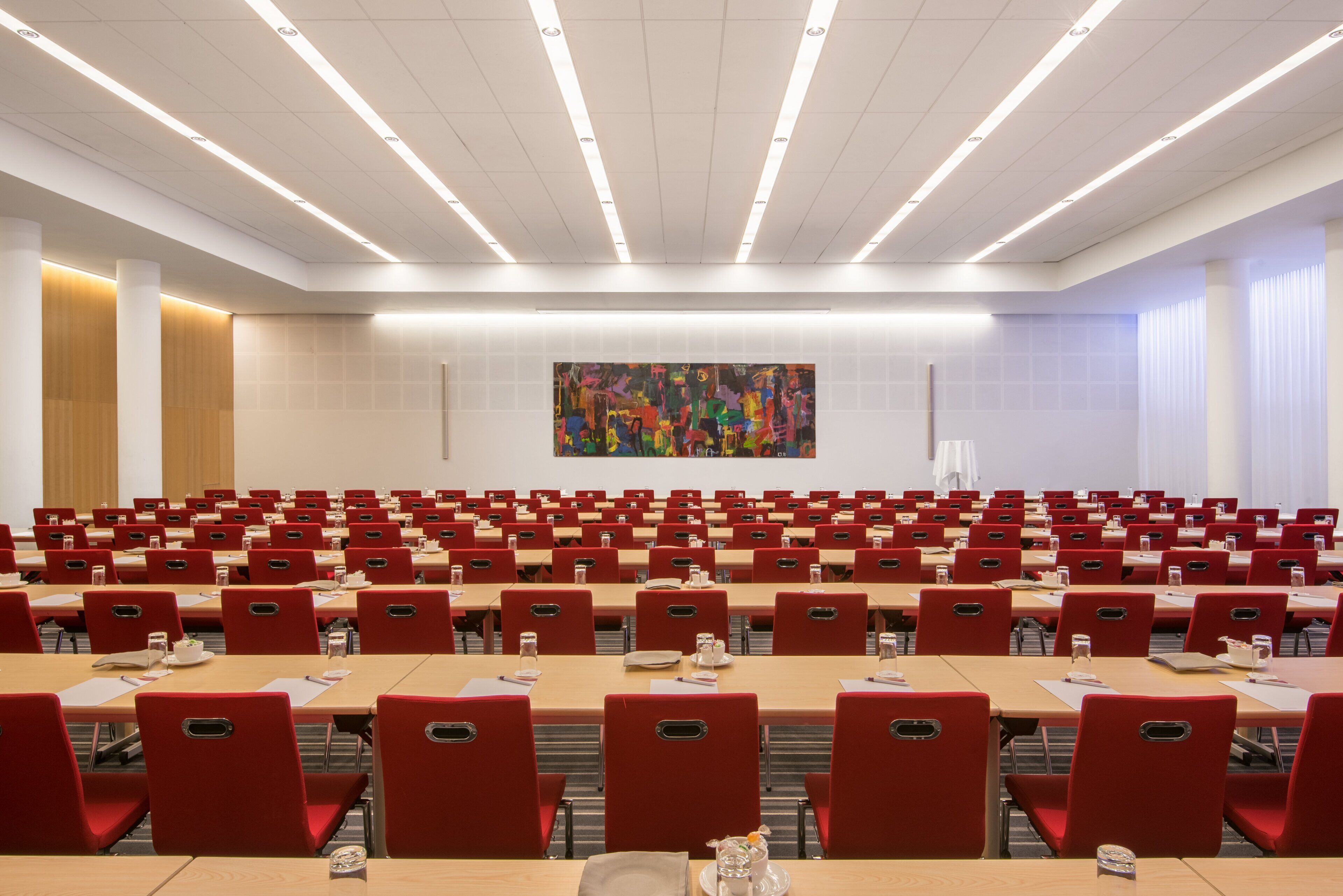 A conference room with rows of red chairs and tables, a large abstract painting on the wall, and a modern ceiling design.