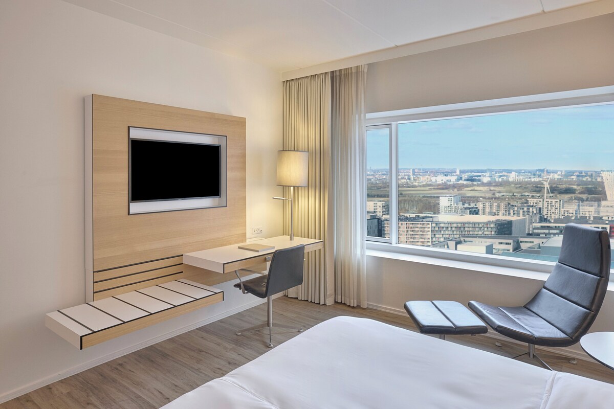 A modern hotel room with a large bed, a desk with a chair, a television mounted on the wall, and a large window offering a city view.