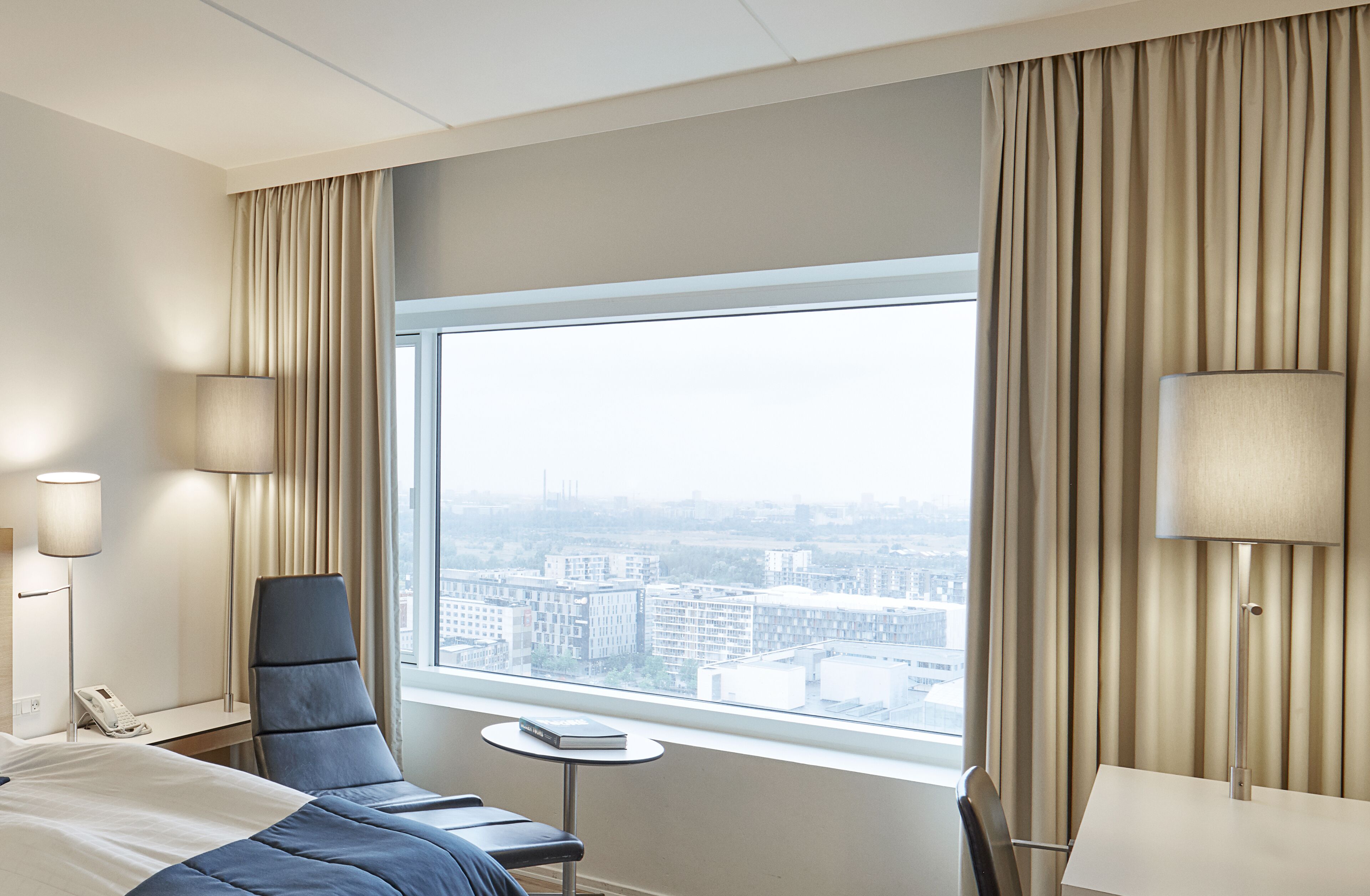 A hotel room with a large window, a bed, a chair, a small table, and a lamp.