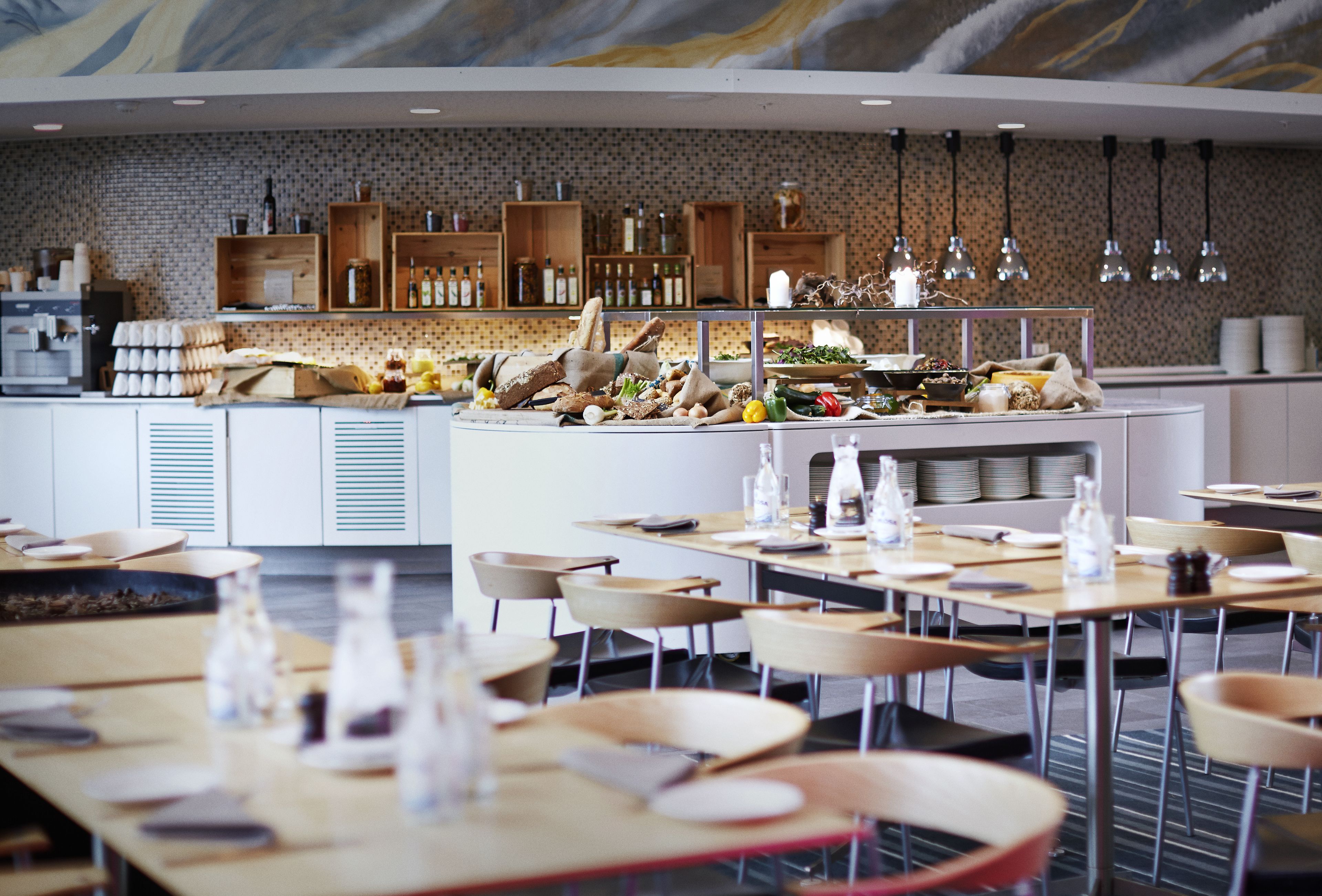 A modern restaurant with wooden tables and chairs, a central island with food and drinks, and a brick wall with shelves of bottles and glasses.