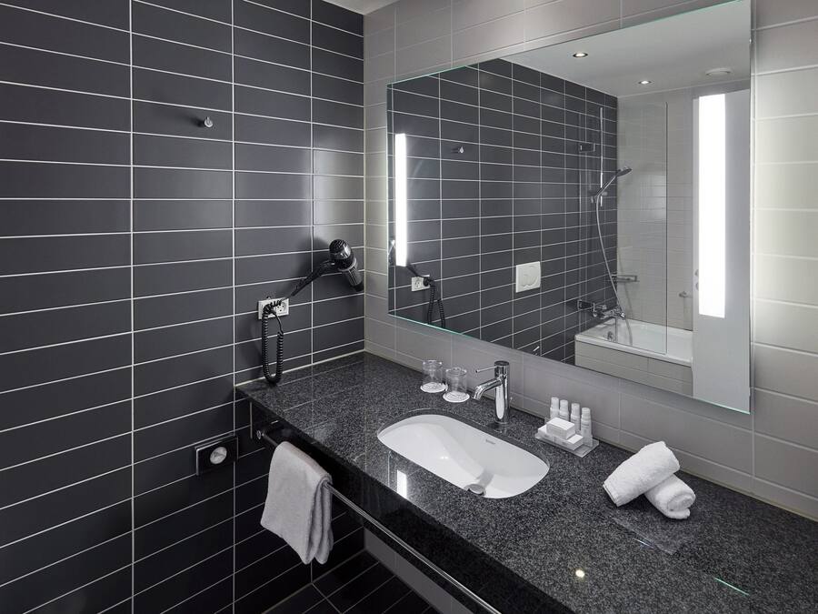 A modern bathroom with black tiled walls, a large mirror, a granite countertop, a white sink, a handheld showerhead, and a towel rack.