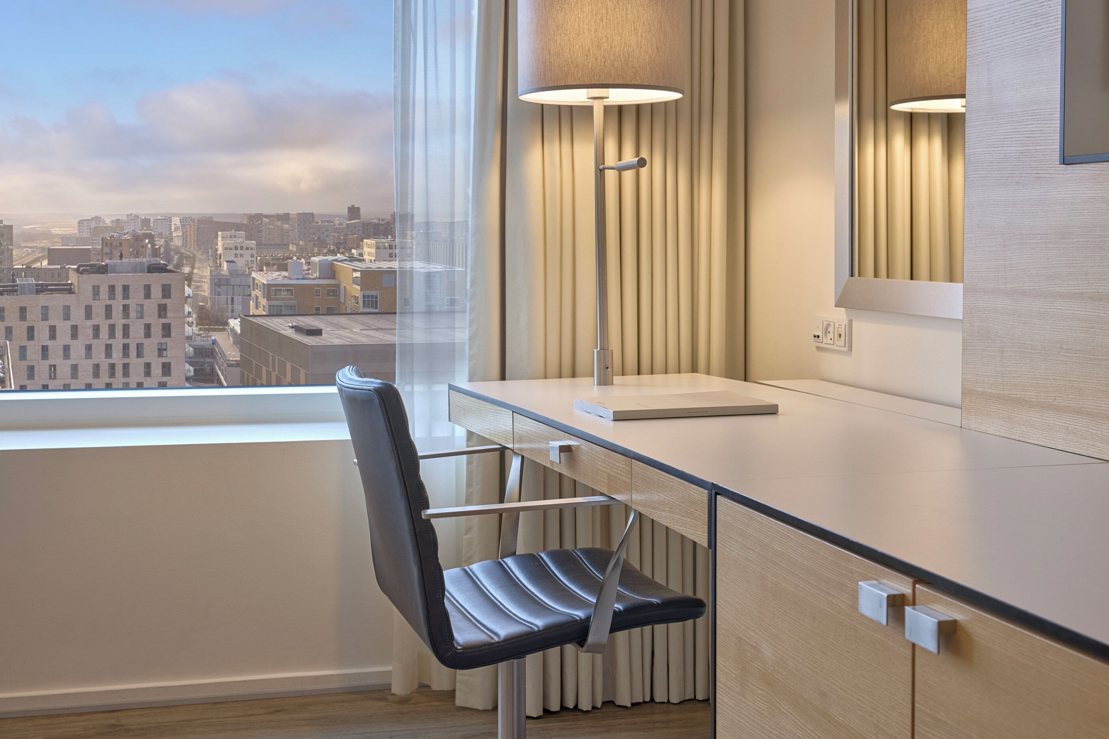 A modern hotel room with a desk, chair, lamp, and a view of the cityscape.