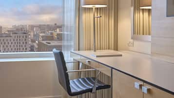 A modern hotel room with a desk, chair, lamp, and a view of the cityscape.