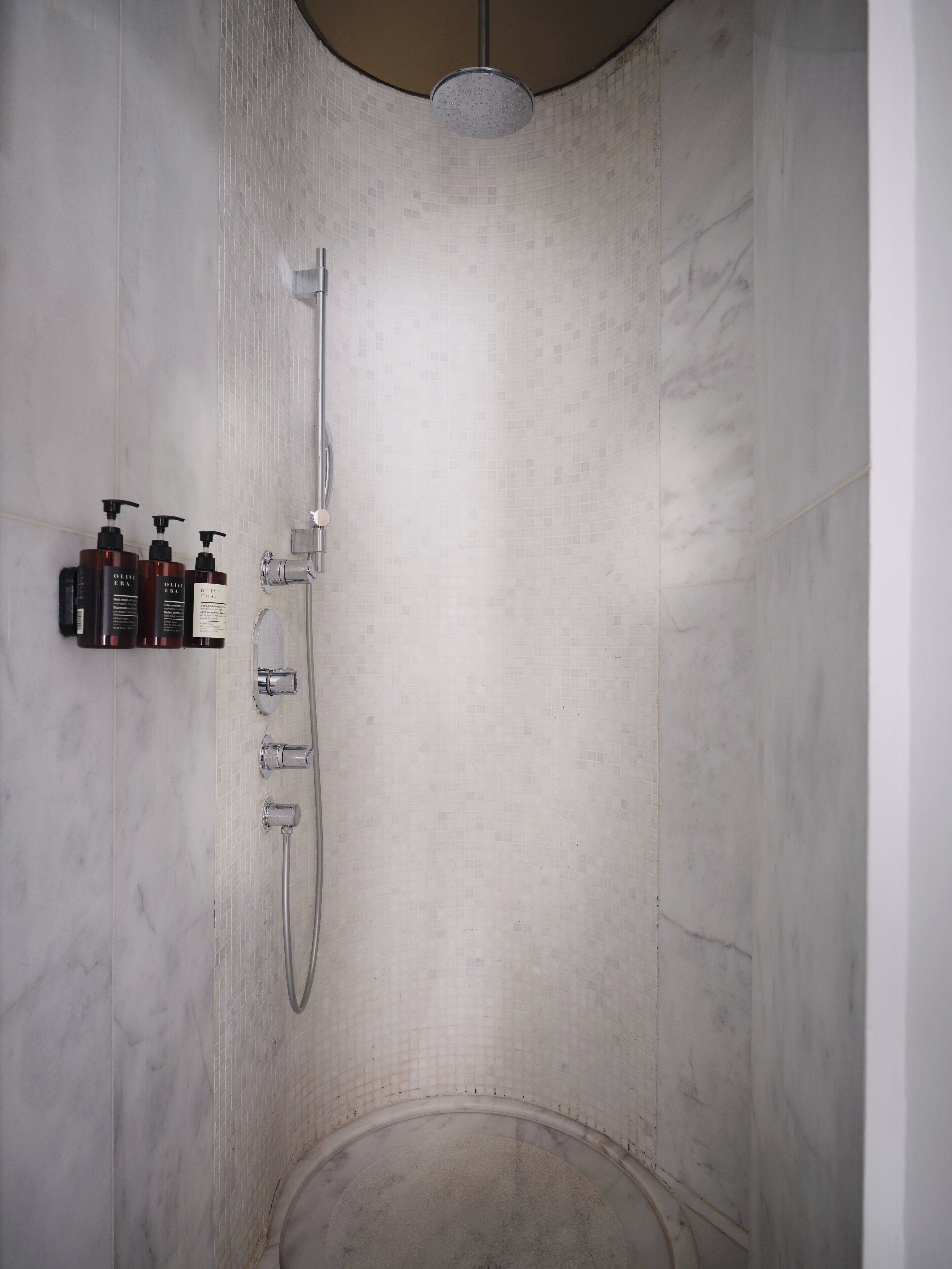 A modern shower with a handheld showerhead and a wall-mounted faucet. There are three bottles of shampoo and conditioner on a shelf to the left.