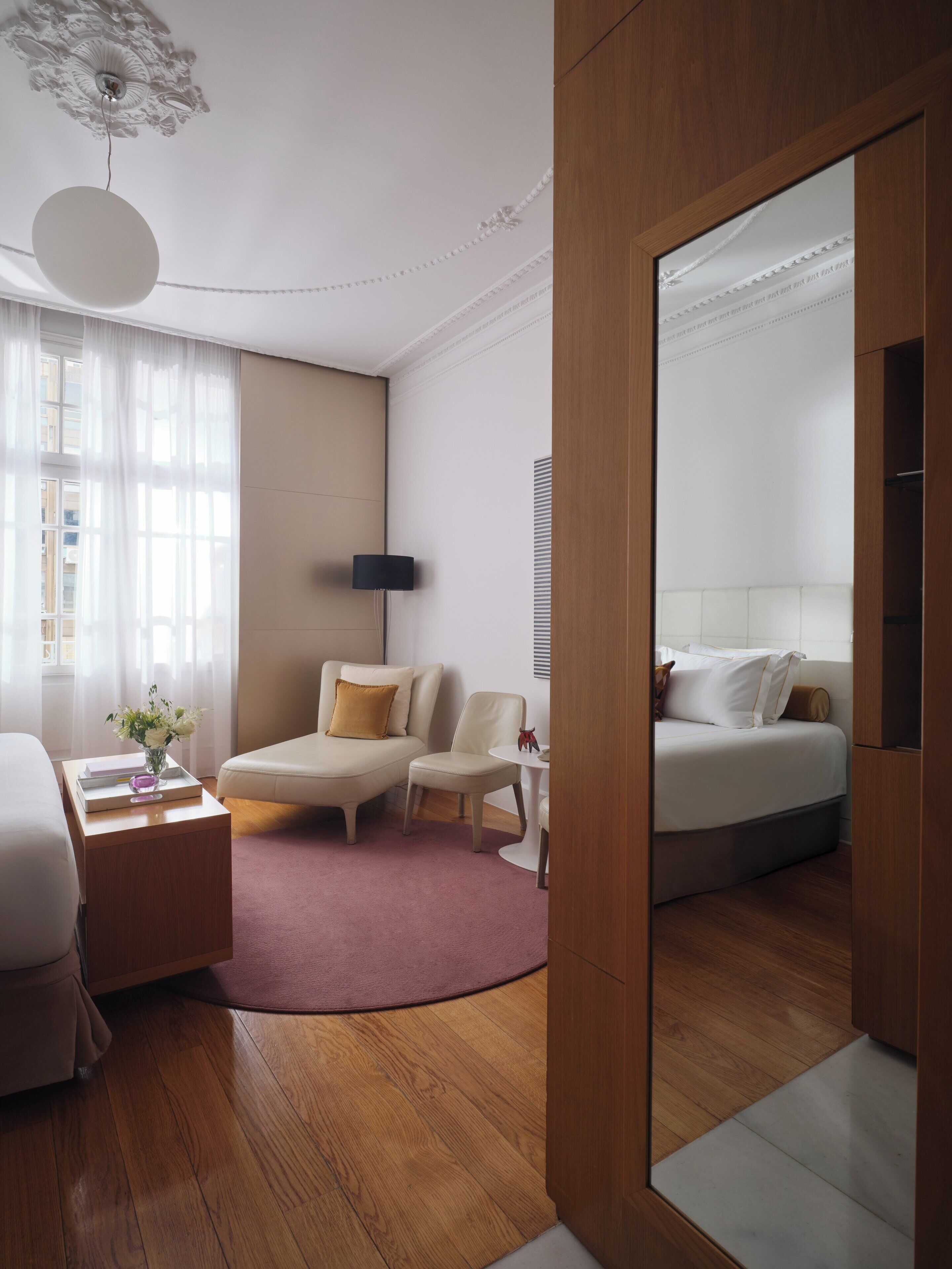 A modern hotel room with a wooden floor, a bed, a sofa, a small table with a lamp, and a large mirror.