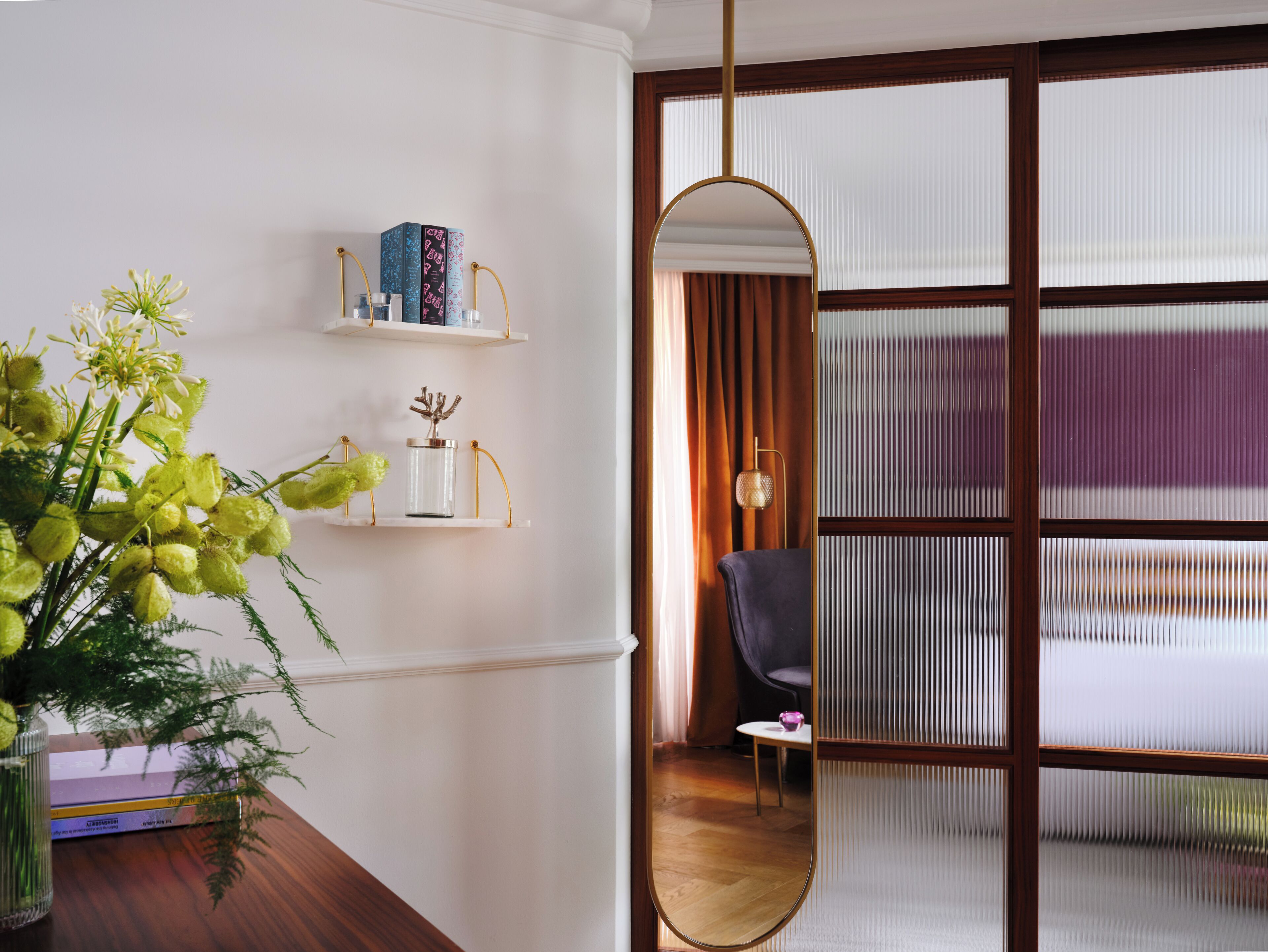 A room with a wooden floor, a mirror, a bookshelf with books, a vase with flowers, and a window with blinds.