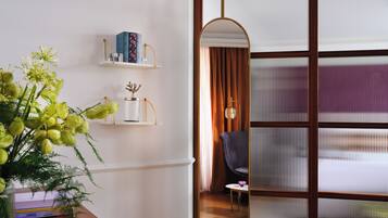 A room with a wooden floor, a mirror, a bookshelf with books, a vase with flowers, and a window with blinds.