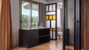 A modern room with a wooden floor, a dark wooden cabinet, a glass table with a bottle and glasses, and a view of greenery outside.