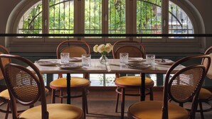 Comedor con mesa dispuesta para seis personas, sillas de mimbre con cojines amarillos, un florero con flores blancas y vistas a la vegetación a través de amplios ventanales de arco.