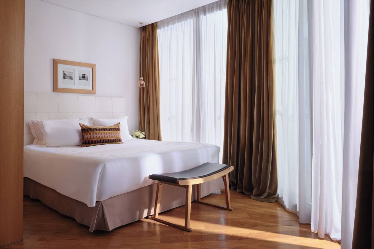 Una cama bien tendida con un banco, un cuadro en la pared y cortinas de encaje.