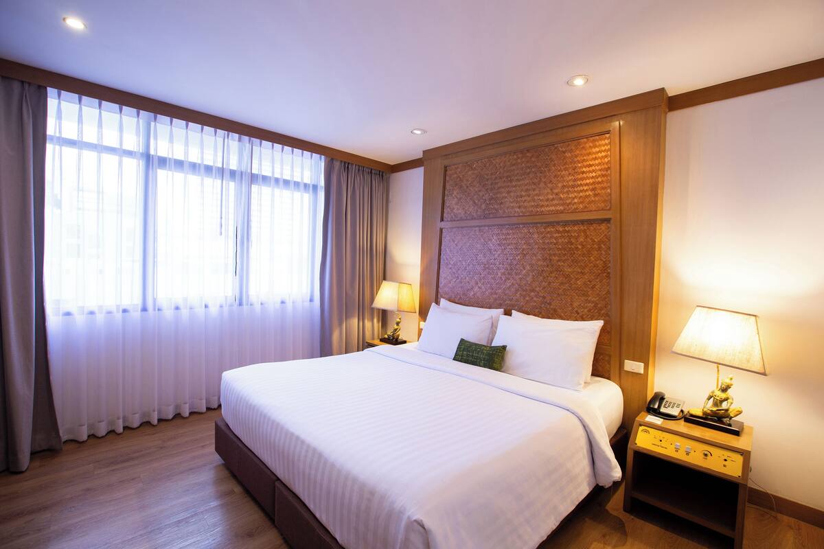 Hotelkamer met een groot bed, twee nachtkastjes met lampen, een houten hoofdbord en een groot raam met gordijnen.