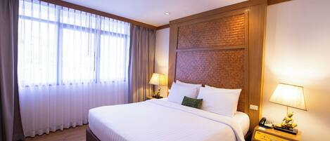 A hotel room with a large bed, two bedside tables with lamps, a wooden headboard, and a large window with curtains.