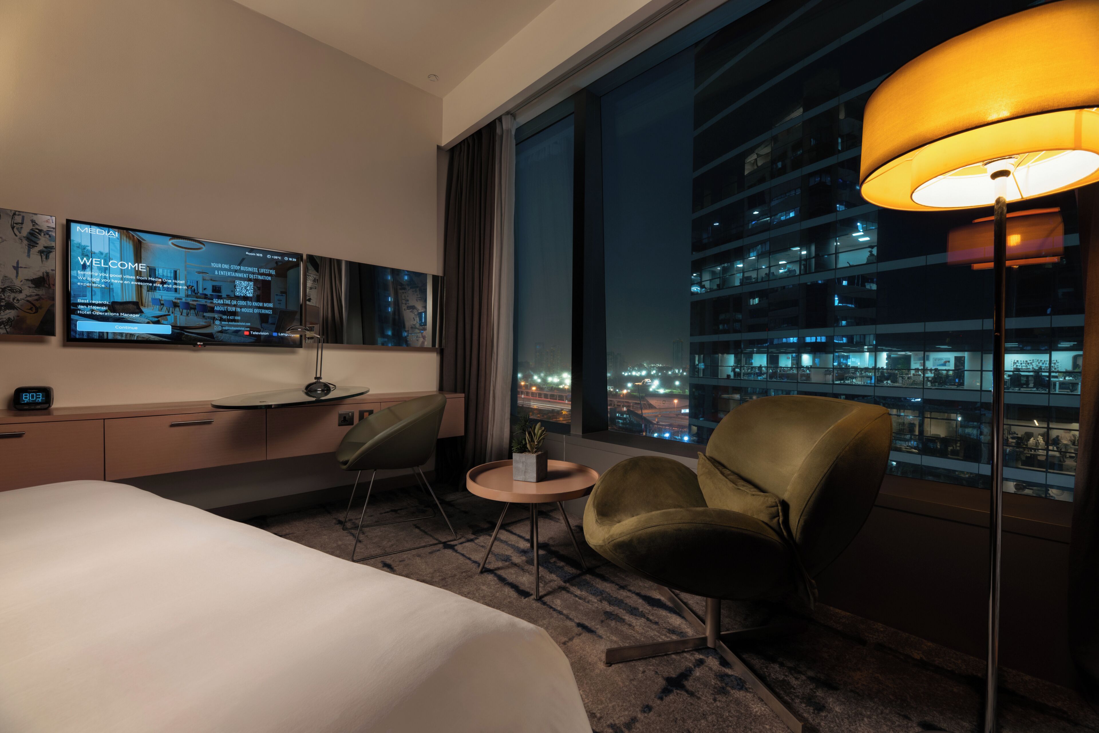 Una habitación de hotel con una cama, un escritorio con una computadora, una silla, una mesita pequeña, una lámpara y un ventanal con vista a la ciudad.