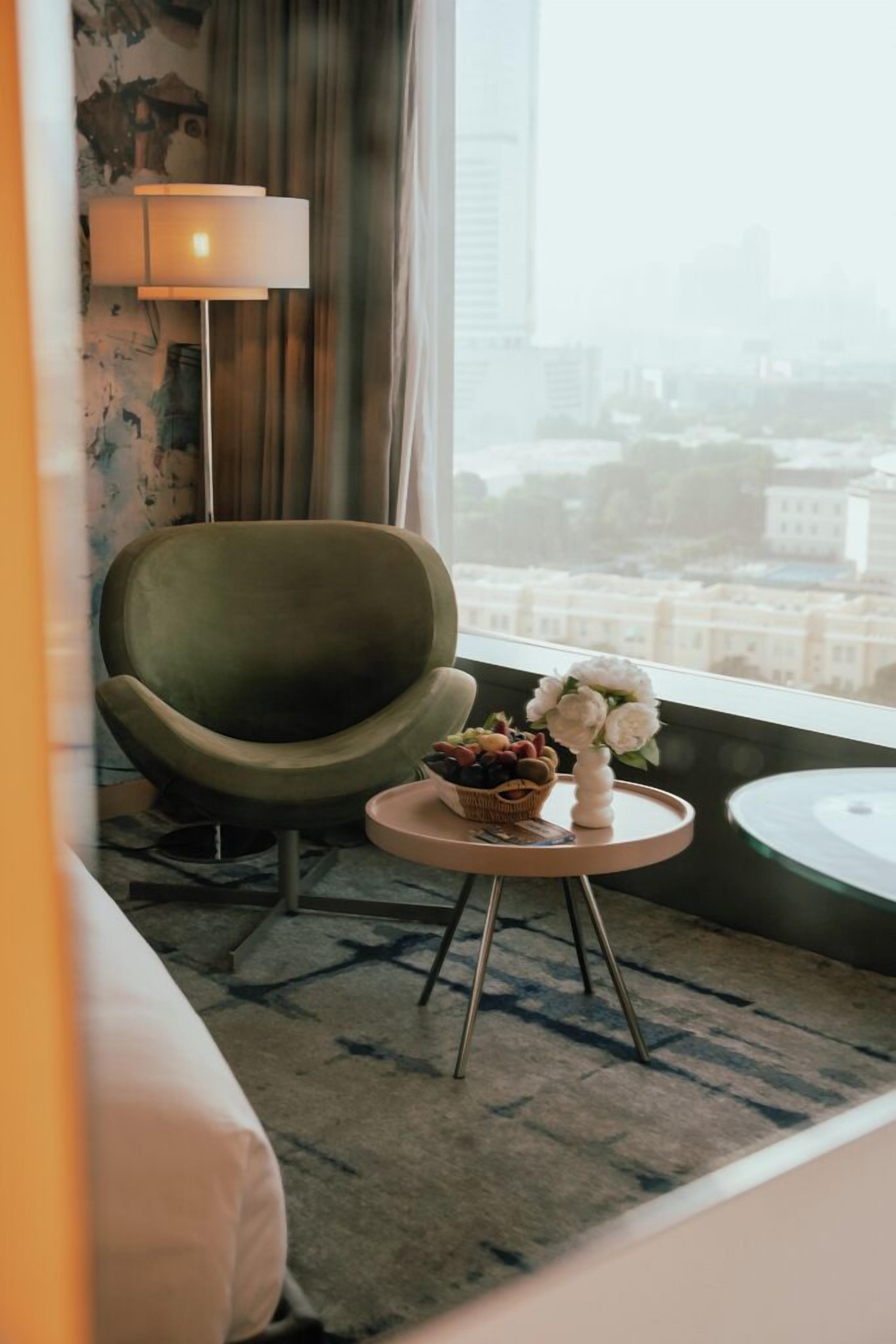A room with a green armchair, a small round table with a basket of fruit and flowers, a lamp, and a view of a cityscape through a window.