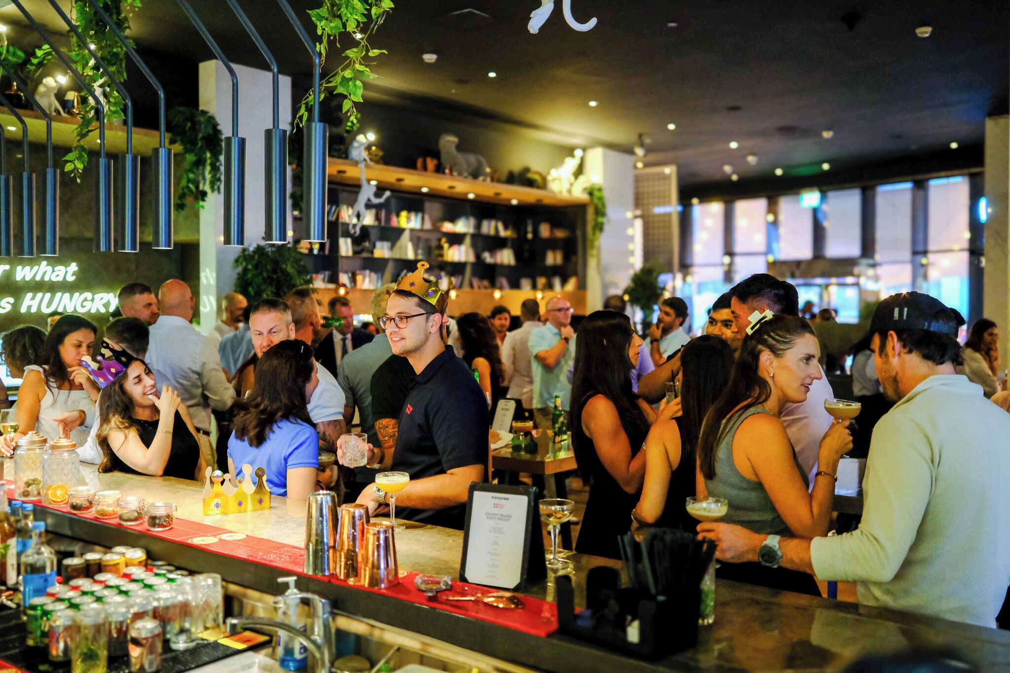 A busy bar area with a bar counter, shelves with bottles, and a menu board.