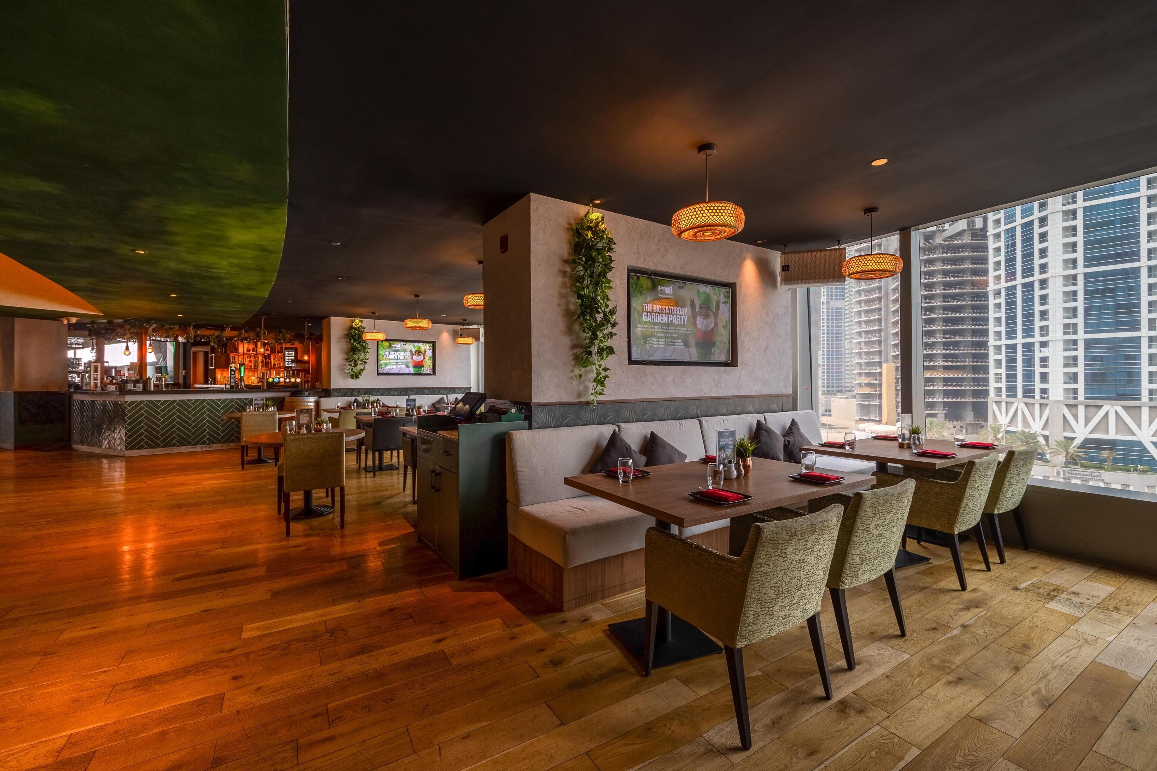 Un restaurante moderno con zona de bar, suelo de madera y grandes ventanales con vistas a la ciudad.
