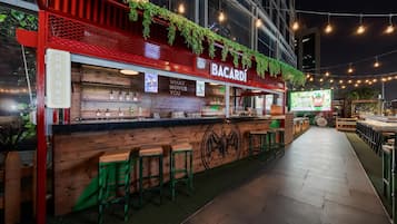 An outdoor bar area with a Bacardi sign, wooden bar counter, green stools, and a pathway leading to more seating.