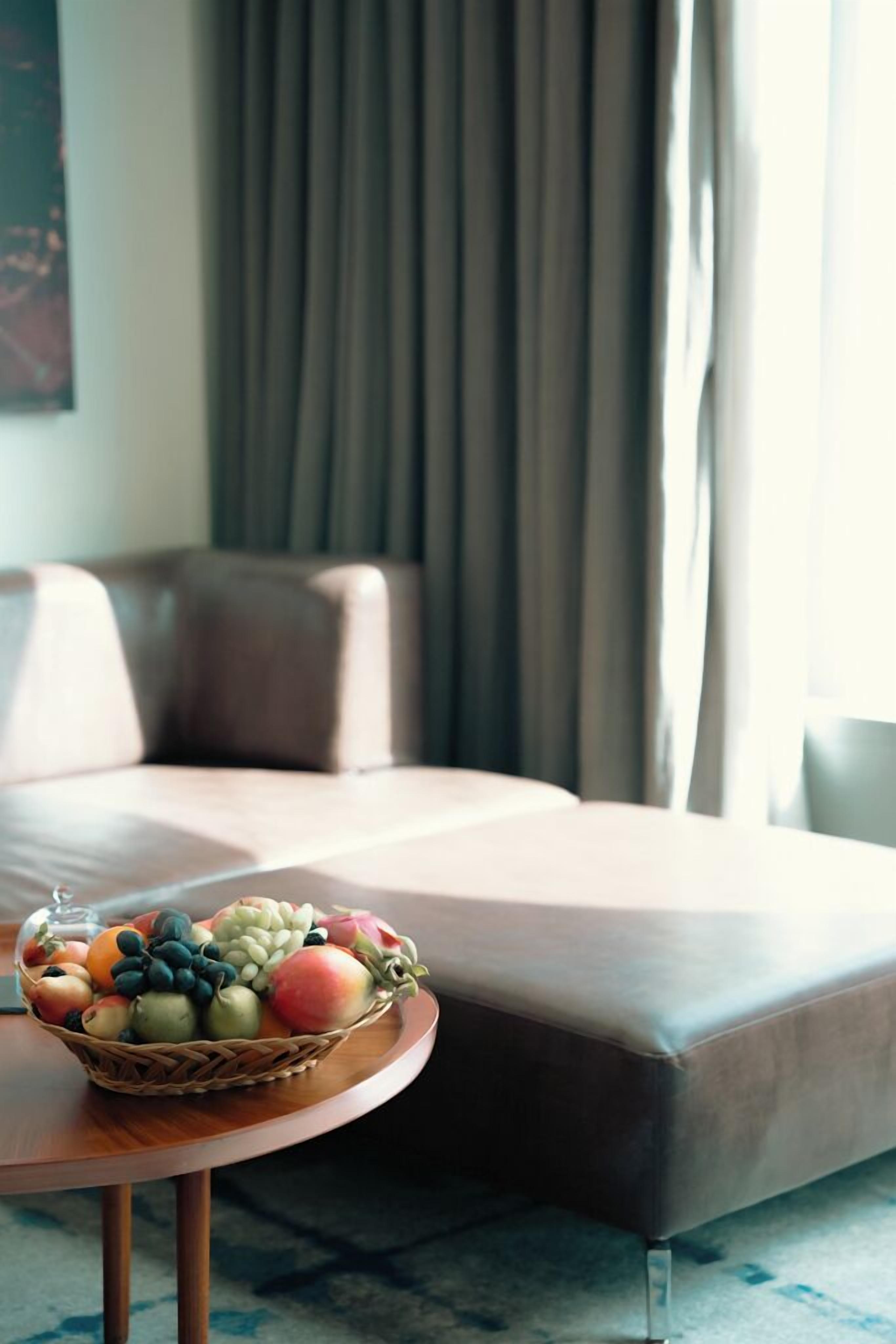 Una sala de estar moderna con un sofá, una mesa redonda con una cesta de fruta y una ventana con cortinas.
