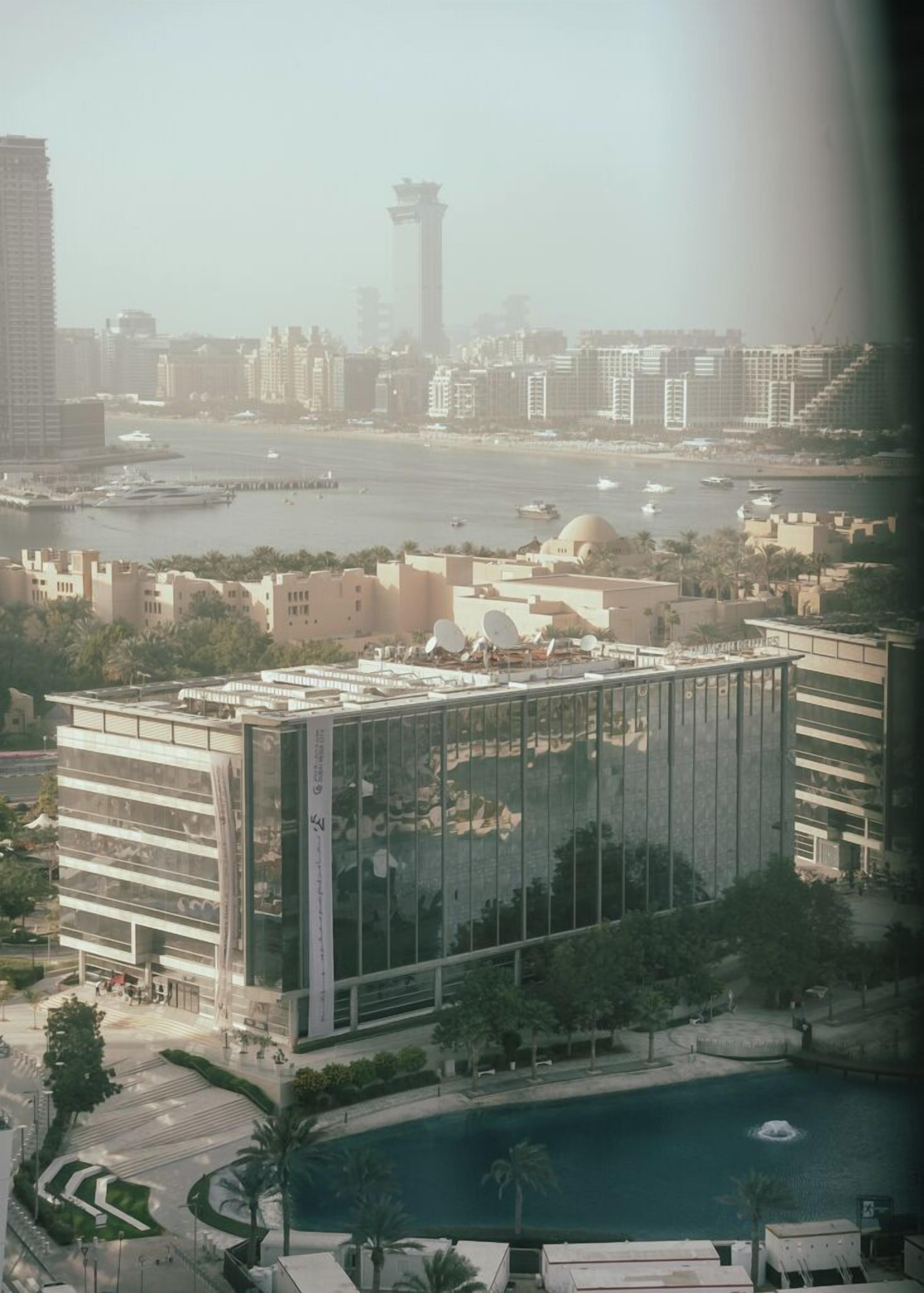 A modern building with a glass facade, a central courtyard, and a view of a body of water with boats.